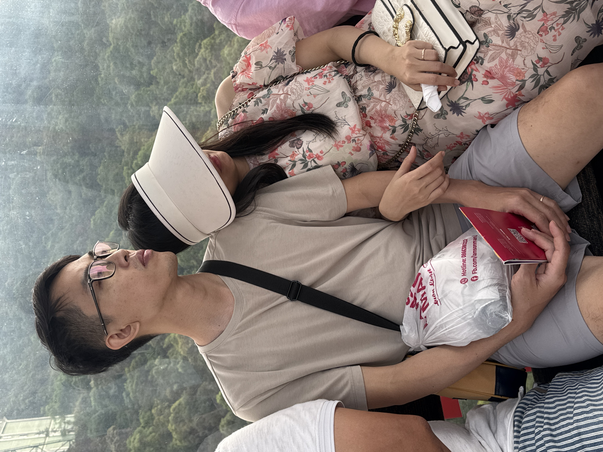 A rotated medium shot shows a seated Asian man and woman in an indoor space, potentially a cable car or enclosed transportation. The man, wearing glasses and a beige t-shirt with a black strap across his chest, looks upward and to the left. He holds a plastic bag and a red booklet. The woman, wearing a floral-patterned top and a white visor, rests her head on the man’s shoulder and holds a white purse. The background consists of blurred greenery, possibly indicative of an elevated location. A portion of another person wearing a striped shirt is visible in the bottom right, and a person with a pink shirt is in the upper left corner. The plastic bag appears to have a Vietnamese address on it.