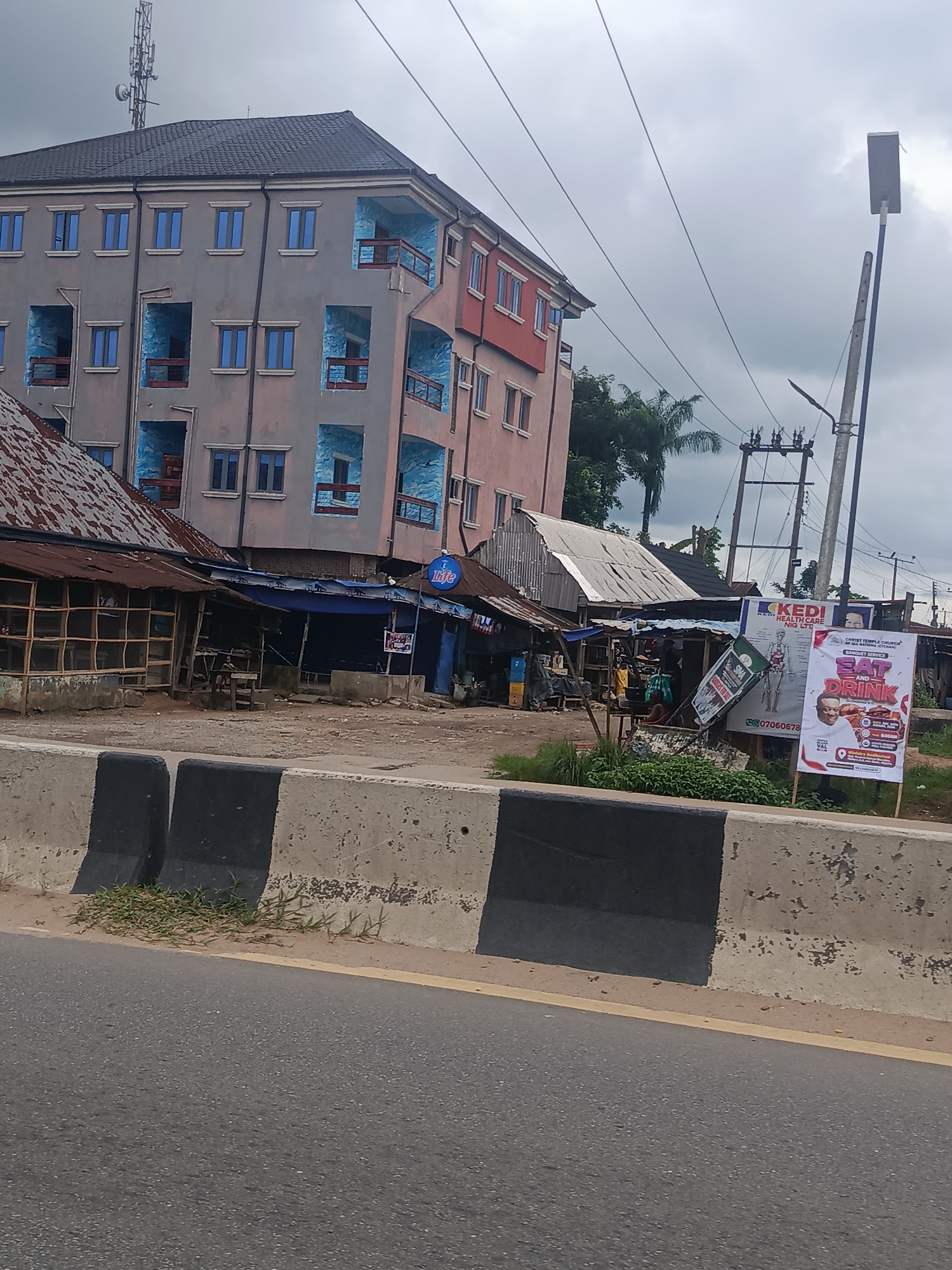 The image depicts a street scene in Mgbidi, Nigeria, characterized by a multi-story building and a collection of smaller structures. The main building, painted in shades of gray and pink, features blue-trimmed balconies and numerous windows. To its left, a row of single-story shops with corrugated metal roofs is visible, some appearing to be open for business. In the foreground, a concrete barrier with black and white stripes separates the road from the scene. Several billboards and signs are displayed, including one for "KEDI HEALTH CARE NIG LTD" and another advertising a "BANQUET SERVICE EAT AND DRINK" by "CHRIST TEMPLE CHURCH". A street light is visible on the right side of the image, and power lines crisscross the sky, which is overcast. The ground is a mix of dirt and pavement. A few individuals are present, primarily near the smaller shops, some seated or standing. The overall impression is of a bustling, though somewhat disheveled, commercial area.