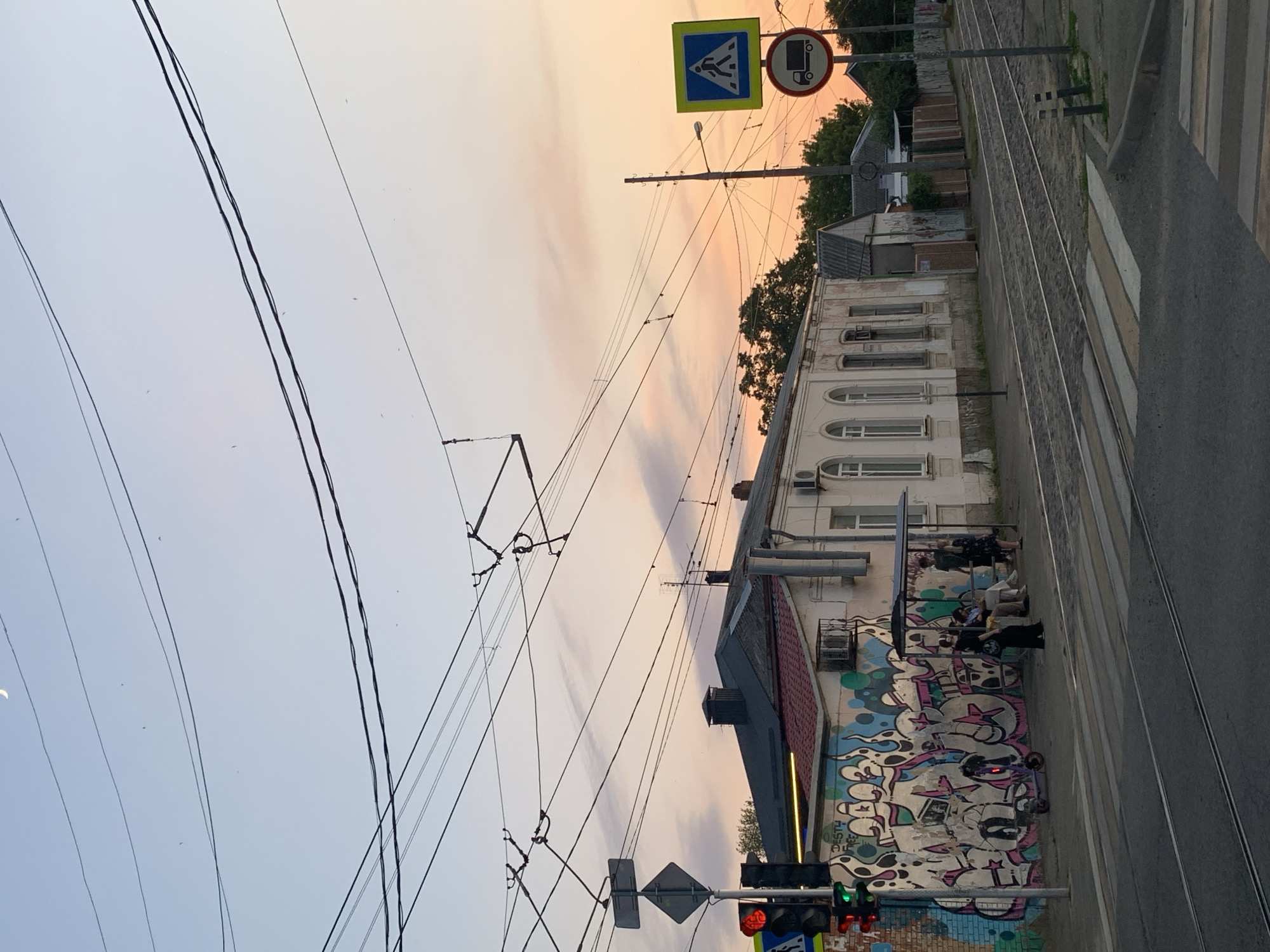 The image depicts a street scene at dusk in an urban environment, possibly in Unknown, Unknown. Overhead power lines, likely for trams or trolleys, crisscross the sky, which is colored with soft hues of orange and pink from the setting sun. A crescent moon is visible in the upper left corner.
In the foreground, tram tracks run across a paved road. A pedestrian crossing is marked by white stripes. To the left, a traffic light shows red on top and green on the bottom. A street sign with a diamond shape is mounted on a pole.
Along the sidewalk, there is a building with a graffiti-covered wall. The graffiti features large, stylized lettering and abstract shapes in various colors, including pink, white, black, and teal. Adjacent to the graffiti is a bus stop shelter where several people are waiting. A person is seated on the bench, and two others are standing. A purple electric scooter is parked near the graffiti wall.
The building behind the graffiti is a long, light-colored structure with arched windows. Further down the street, more buildings and trees are visible, along with street signs. One sign is a blue square with a white pedestrian symbol, and below it, a circular sign with a red border and a black truck symbol. The overall atmosphere is calm and quiet, suggesting the end of the day.