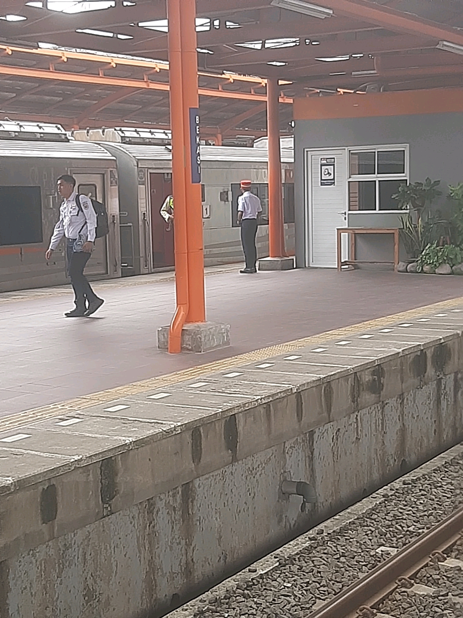 The image depicts a scene at a train station in Bekasi, Indonesia, specifically at the "BEKASI" station as indicated by signage on a prominent orange pillar. The foreground is dominated by a concrete train platform edge, with textured yellow tactile paving lines. Beyond the platform, a train is visible, with its doors open. Two individuals are present. One is a man wearing a white shirt, dark pants, and carrying a black backpack, walking on the platform. Another individual, wearing a light-colored shirt and a red hat, stands further down the platform near an open train door. In the background, a building with a grey facade features a white door and a window. Plants are visible to the right of the building. The station's roof structure, with orange beams and white fluorescent lighting, is visible overhead. The overall environment suggests an active, open-air train station.