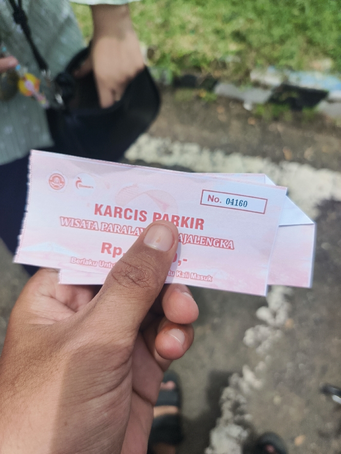 A person's hand is holding a pink parking ticket with the words "KARCIS PARKIR" and "WISATA PARALAYAN JALENGKA" printed on it. The ticket number is "04160". The ticket appears to have been folded in half. The background of the image shows blurred, out of focus details of a person and a paved walkway. The coordinates provided are near the location of the "Wisata Paralayang JALENGKA" in Majalengka, Indonesia, indicating the ticket likely belongs to the paragliding site.