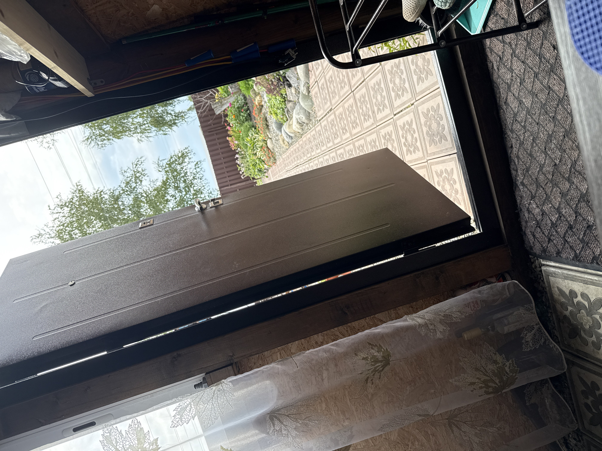 The image shows an open doorway leading to an outdoor garden area in Krasnogorskoye, Russia. Inside the structure, a dark brown metal door is ajar, revealing a textured wooden wall and a sheer white curtain with a leaf pattern hanging to the left. A bottle is partially visible behind the curtain. To the right of the doorway, a black metal shoe rack is positioned, with a shoe and a light blue object on it. Beyond the doorway, a paved pathway with patterned tiles leads to a colorful garden filled with flowers and rocks, bordered by a brown wooden fence and mature trees. The sky above is partly cloudy. The scene suggests a residential or communal outdoor space.