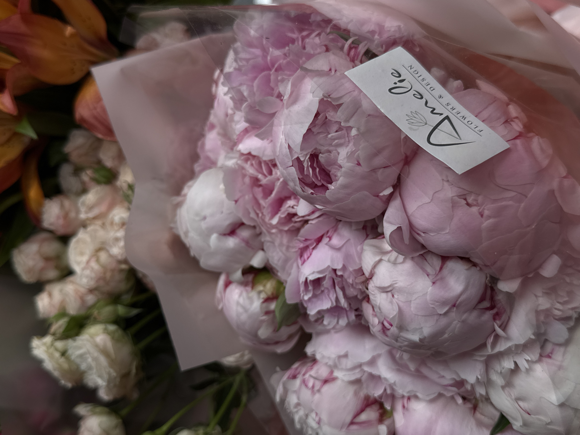 A close-up shot shows a bouquet of pink peonies, prominently displayed. A white tag with the text "Amelie FLOWERS & DESIGN" is affixed to the bouquet. Additional flowers, including orange alstroemeria and small white roses, are also visible in the image, though less in focus. The location is Yerevan, Armenia, inferred from the tag potentially indicating a local flower shop. No people are visible.