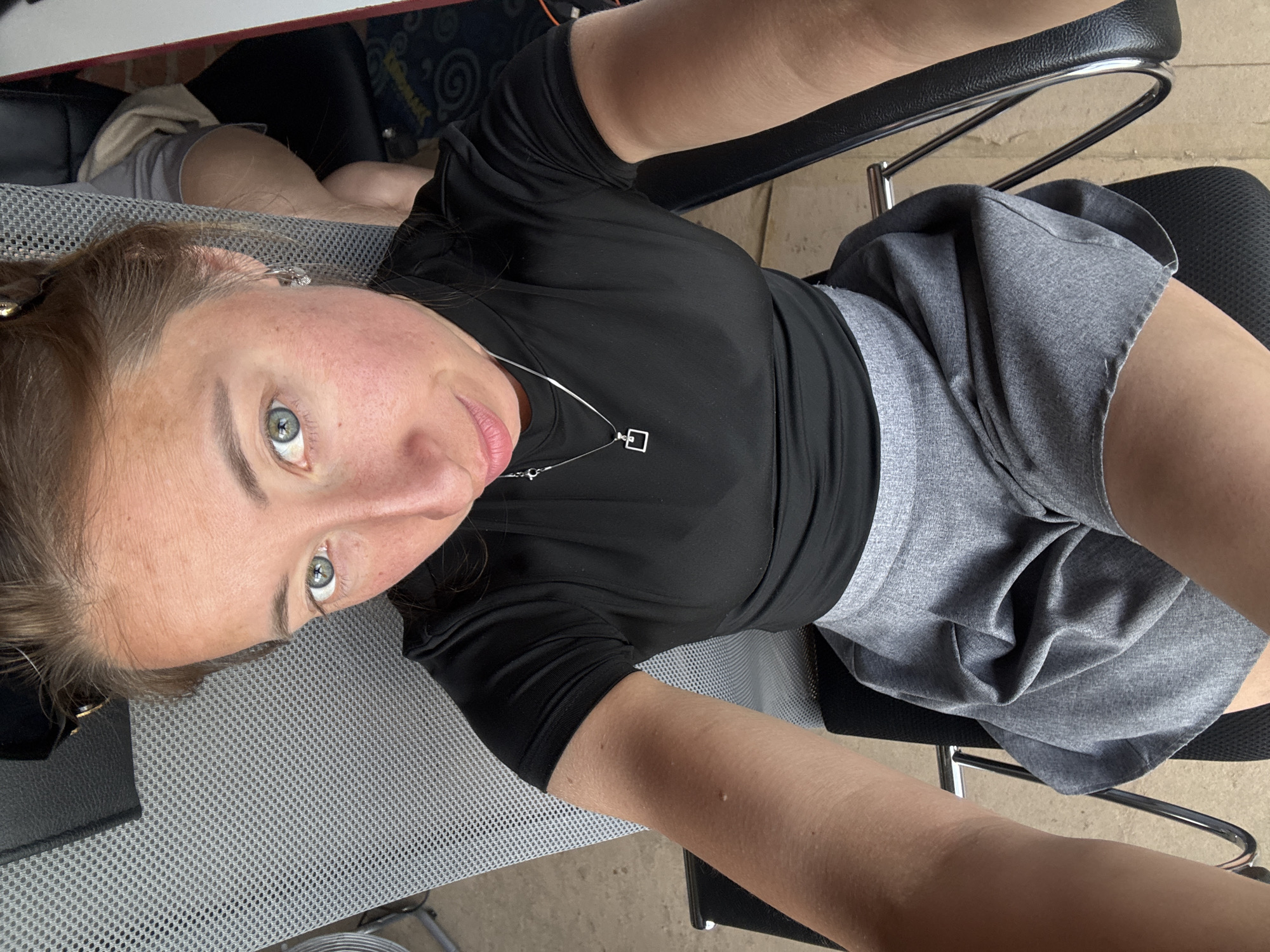 A woman with light skin and blue eyes is looking directly at the camera. She is wearing a black shirt and a gray skirt. She is sitting in a gray office chair. A silver necklace with a square pendant is visible on her chest. The environment appears to be indoors. There is no information to confirm this image was taken in Chaikovsky, Russia.