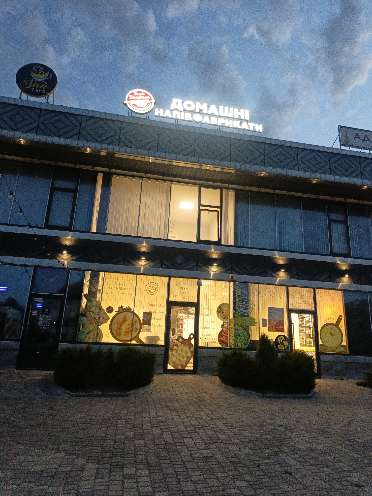 This is a medium-angle, eye-level shot of a modern building facade at dusk. The building has multiple stories with large windows, some of which are illuminated from within, casting a warm glow on the exterior. The facade is decorated with patterned metalwork above the ground-floor windows.
Above the building, signs are visible against the twilight sky, which is filled with dramatic clouds. One sign reads "Sun CAFE" with a stylized coffee cup logo. Another illuminated sign, in a rounded shape, contains Ukrainian text. A larger, illuminated sign across the top of the building prominently displays "ДОМАШНІ НАПІВФАБРИКАТИ," which translates to "Homemade Semi-Finished Products." A partial sign for "АД" is also visible on the right.
The ground floor features large display windows with illuminated graphics depicting food items and text in Ukrainian, such as "Готуємо по-домашньому" (We cook homemade) and a list of dishes like "Вареники" (Varenyky), "Пельмені" (Pelmeni), and "Сирники" (Syrnyky). The entrance to the "Sun Cafe" is visible on the left, with operating hours displayed: "9:00-20:00" and "10:00-20:00".
In front of the building, a paved walkway leads to the entrance, with several neatly trimmed evergreen bushes placed strategically. The overall impression is of a commercial establishment, likely a cafe and a shop selling homemade food items, operating in the early evening. The lighting and the sky suggest the transition from day to night.