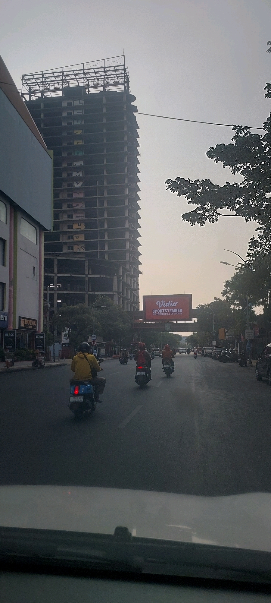 The image shows a view of a street in Surabaya, Indonesia, with a tall, unfinished building in the background. The building appears to be under construction, with exposed concrete floors and scaffolding at the top. On the side of the building, large colorful letters are visible, possibly part of graffiti or an unfinished advertisement.
In the foreground, a road is visible with several motorcycles and their riders. One rider in a yellow jacket is prominently positioned in the center, on a blue scooter. Other riders are also on motorcycles, heading in the same direction. On the right side of the street, several parked motorcycles and cars are visible.
A large red billboard advertising "Vidio SPORTSTEMBER SEPTEMBER FULL OF SPORTS" is suspended above the road in the middle ground. Trees line the sides of the road, and streetlights are also present. The sky is overcast, suggesting it might be morning or late afternoon. The overall scene depicts a typical urban streetscape in Surabaya, with ongoing construction and daily traffic.