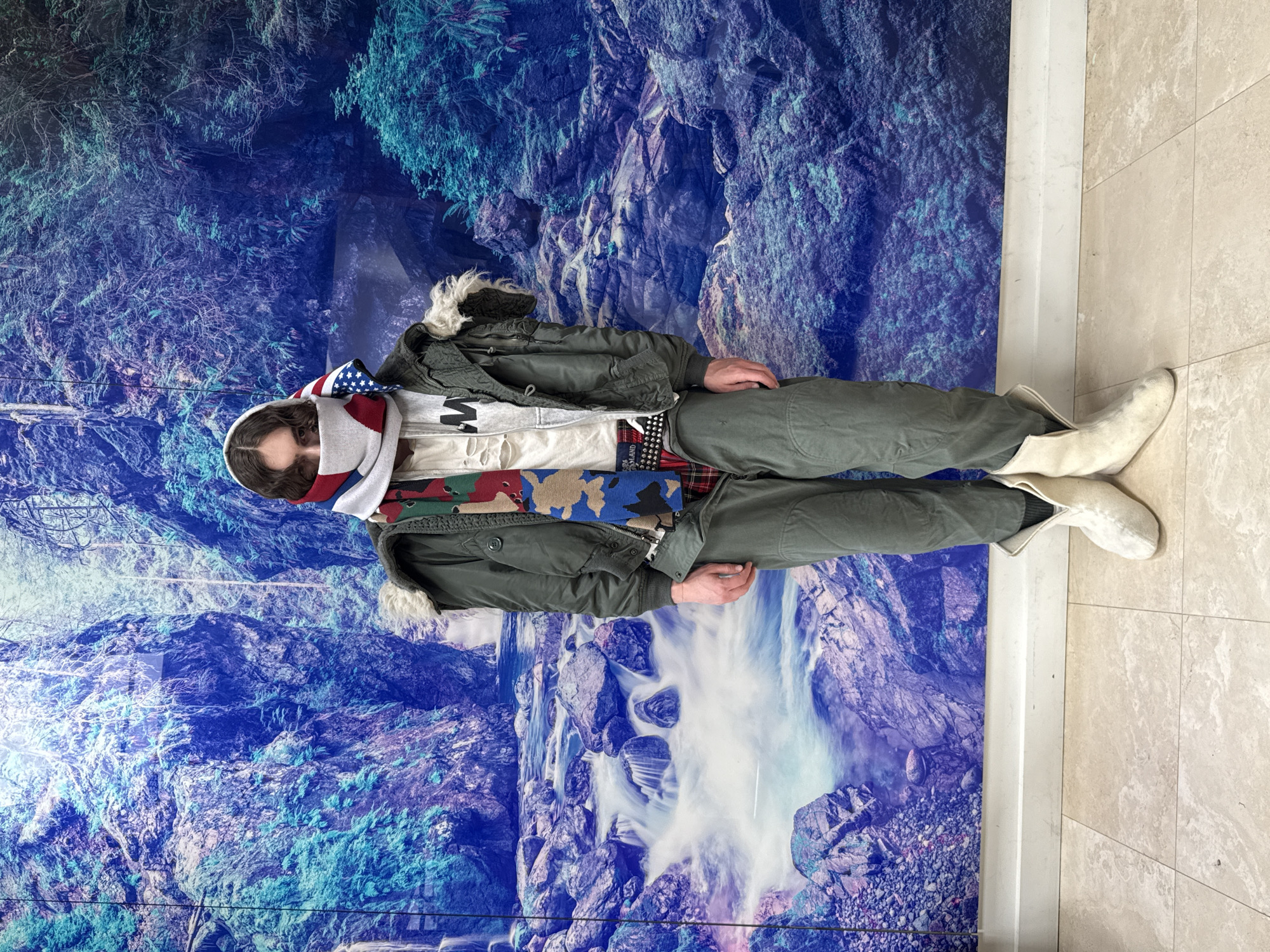 A person stands indoors against a wall decorated with a vibrant, blue-toned nature scene featuring mountains and a waterfall. The individual is fully clothed, wearing an olive green jacket with a fur-trimmed hood, layered over a gray t-shirt with a black "M" graphic and several scarves, one featuring an American flag design. They are also wearing olive green pants with a studded belt and white boots. The background wall appears to be a large, decorative mural. The floor is tiled in beige and tan. The location is indoors, in the city of Unknown, Unknown.
