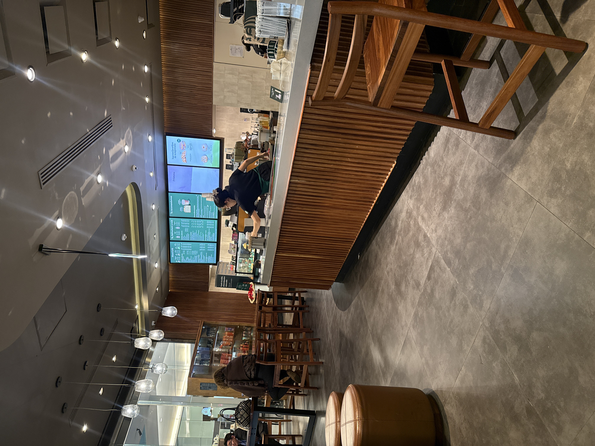 The image captures an interior view of what appears to be a Starbucks coffee shop in Puebla, Mexico. The space features a modern design with wooden paneling on the bar counter and seating area. A barista, dressed in a dark uniform with a green apron, is working behind the counter, possibly preparing drinks. Menu boards displaying product names and prices are visible above the counter. A group of customers is seated in the foreground, and there are additional seating arrangements with wooden chairs in the mid-ground. The lighting is a mix of natural light coming through the storefront windows and pendant lights hanging from the ceiling. The floor is tiled in a neutral color, and the overall ambiance is modern and well-lit.
