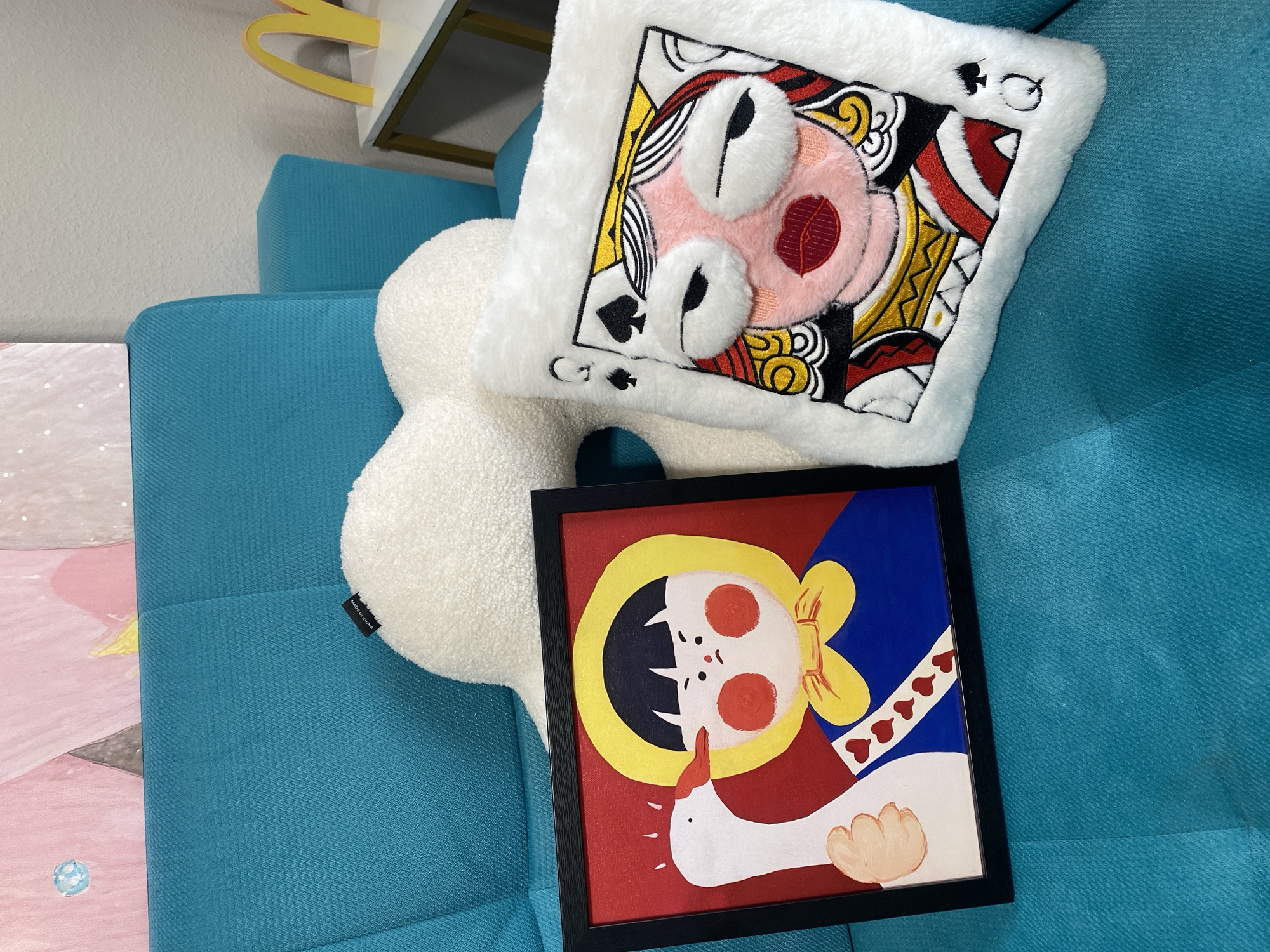 The image shows a close-up of a turquoise-colored sofa. On the sofa, there are two decorative items: a framed artwork on the left and a fluffy white pillow on the right. The framed artwork depicts a stylized illustration of a child wearing a yellow bonnet and blue dress, with a white goose near its face. The goose appears to be pecking at the child's cheek. The artwork has a red background and a black frame. The pillow on the right is white and fluffy, and it features an embroidered design of a queen of spades playing card. The queen's face is depicted with plush fabric elements for the eyes and lips. A white, cloud-shaped pillow is partially visible behind the framed artwork and the queen pillow. In the background, a hint of a pink and abstract artwork can be seen, and to the right, a yellow McDonald's arch logo is visible on a white table. The scene does not provide any indication of a specific location in Moscow, Russia.