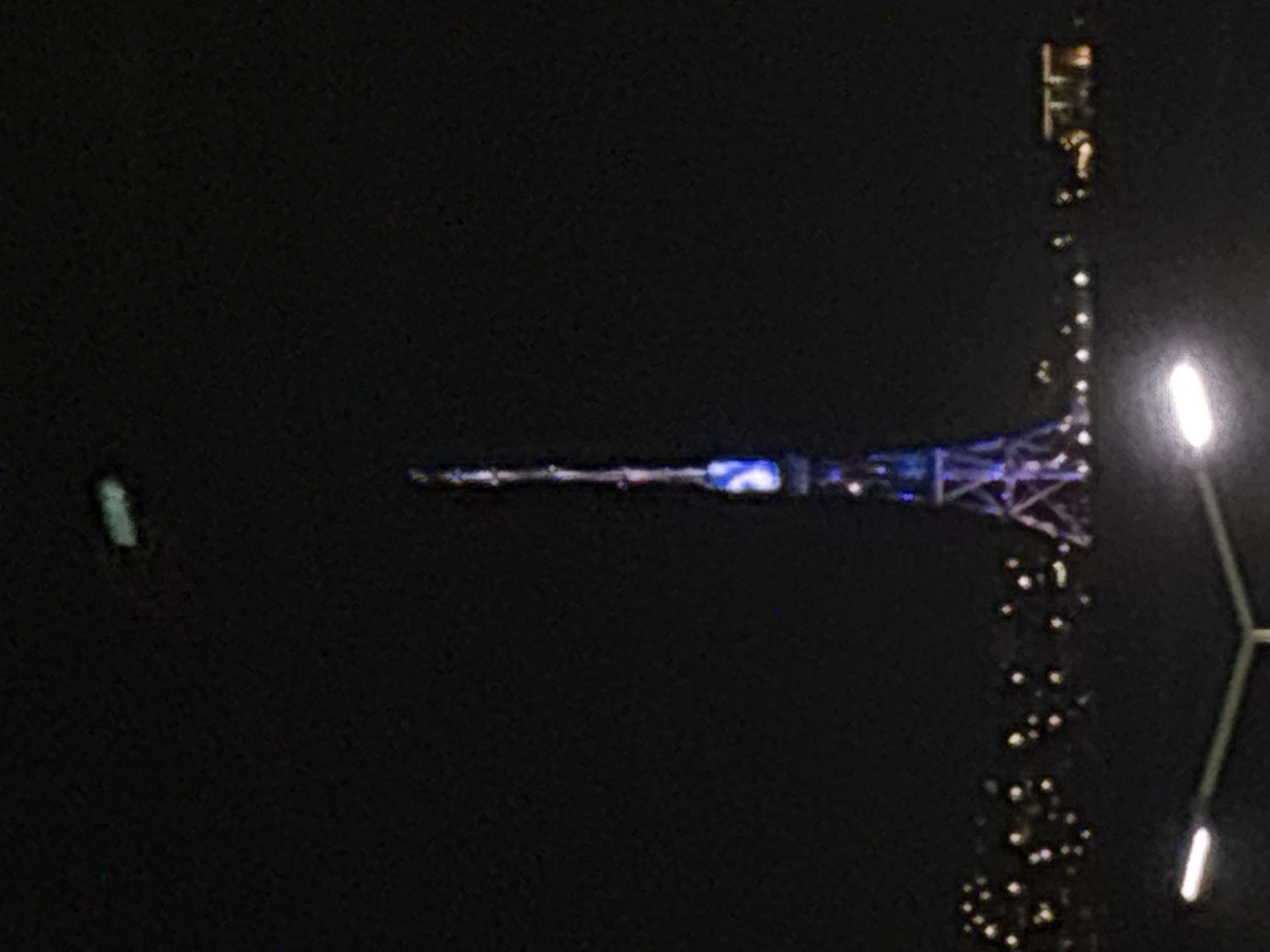 The image depicts a nighttime scene in Yerevan, Armenia, featuring the Yerevan TV Tower illuminated in blue and white light. The tower is viewed horizontally from a distance. To the right of the tower, a line of streetlights stretches towards the foreground, reflecting in what appears to be a body of water. A street lamp with two lights is visible in the bottom right corner. The overall environment is dark, with the lights providing the only sources of illumination.
