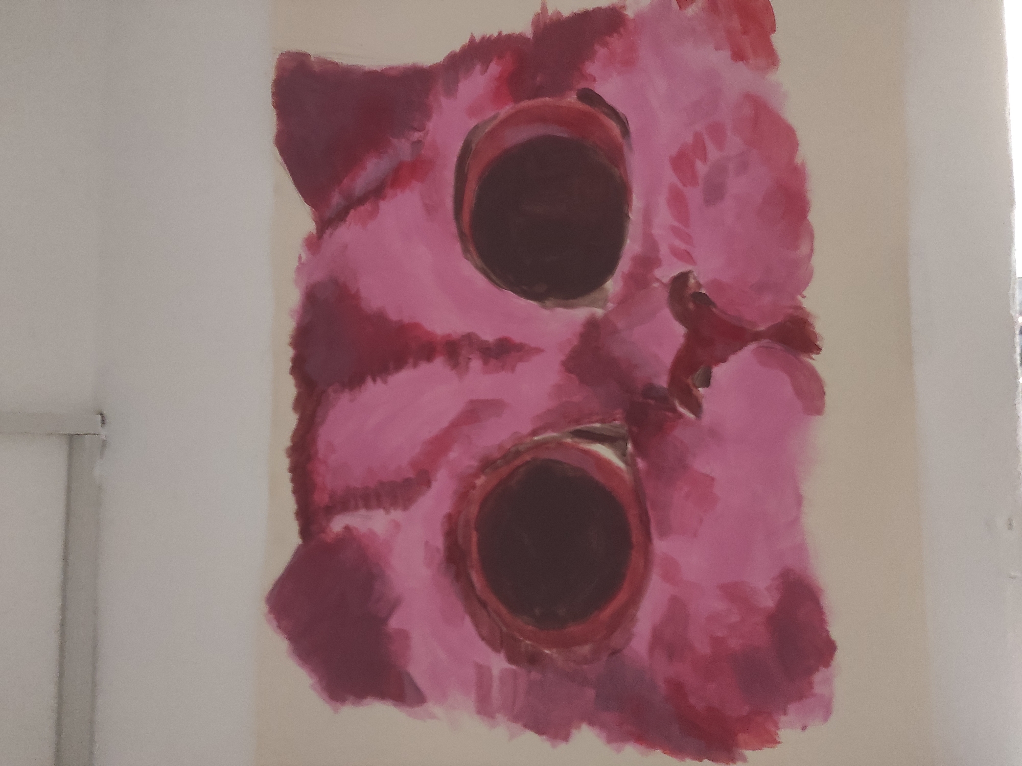 A painting of a pink cat face with dark, circular eyes is on a light-colored wall. The painting is abstract, with rough brushstrokes and undefined edges. The cat has an ear on the left side and a nose on the right side. The painting appears to be indoors near a window, but a city or geographical location is not determinable.