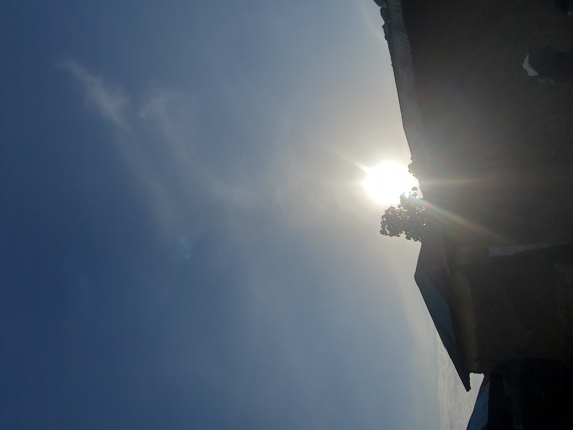 The image shows a low-angle shot of several silhouetted buildings against a bright sky. The sun is setting or rising, creating a strong lens flare that obscures details of the sky and the upper portions of the buildings. The buildings appear to be constructed from mud or rough materials, with corrugated metal roofs. Some foliage is visible in the foreground, silhouetted against the bright sky. The scene suggests a rural or semi-urban setting in Ampan, Nigeria.