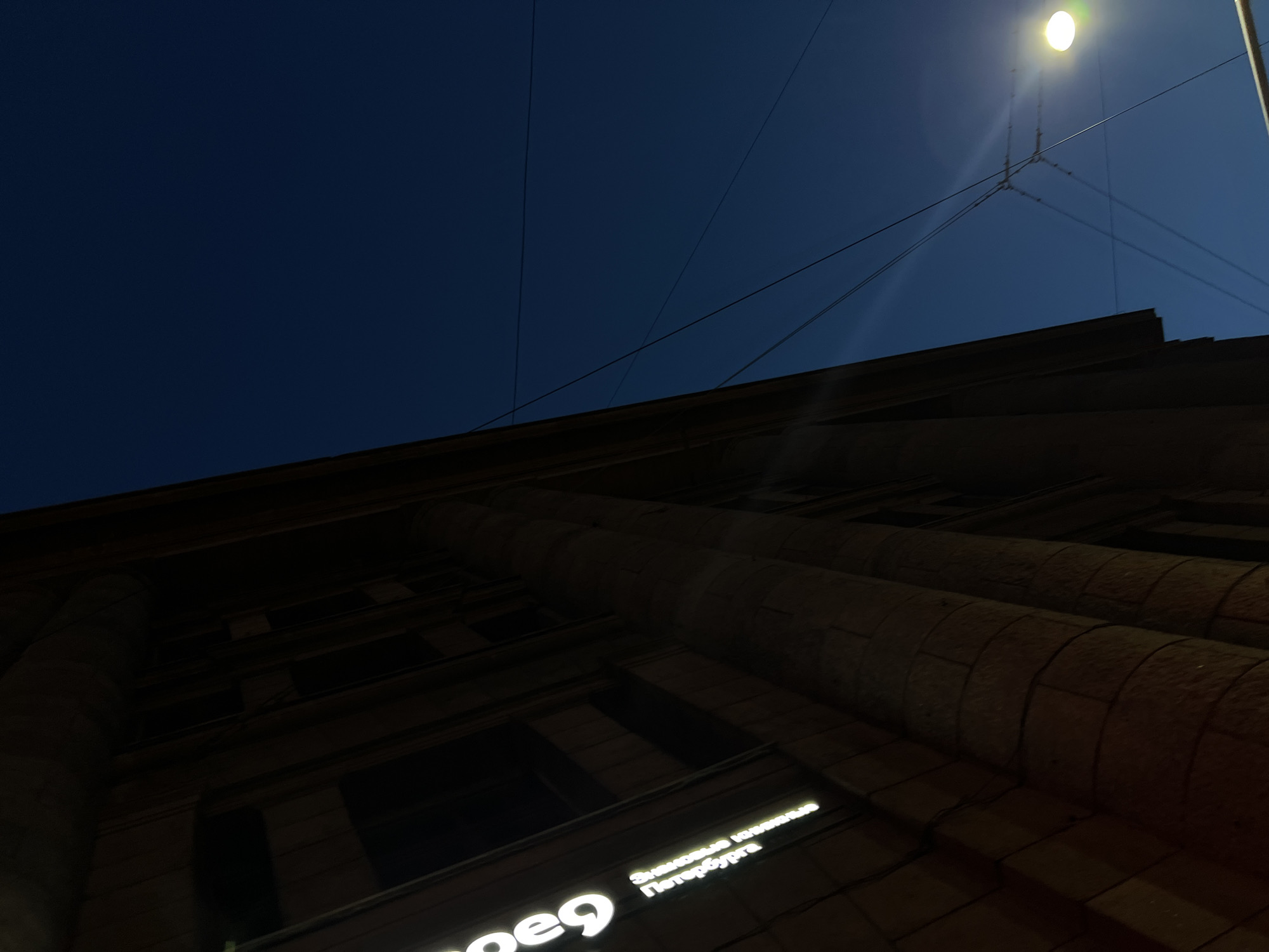 This image captures a nighttime view of a building facade and the dark blue sky in Saint Petersburg, Russia. The building appears to be made of stone with large, rounded columns. A neon sign with white letters reading "peg" and "Знаковые клиенты Петербурга" (Iconic Clients of Petersburg) is visible on the left side of the building. Overhead, electrical wires crisscross the sky, and a bright, round streetlamp illuminates the scene from the right, casting a glow on the wires. The overall impression is one of an urban environment at dusk or night.