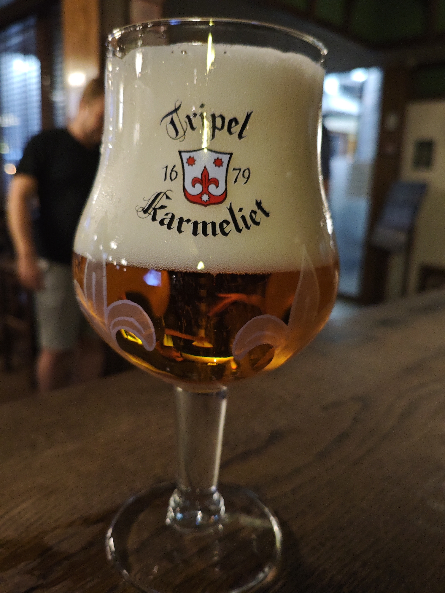 A close-up, eye-level shot focuses on a stemmed glass of beer, brimming with a frothy head. The beer itself is a rich amber color, with subtle reflections visible on the glass's surface. The glass is decorated with black lettering that reads "Tripel Karmeliet" along with a crest featuring a fleur-de-lis and the numbers "16" and "79". The glass rests on a wooden table, which is slightly out of focus. In the blurred background, a person wearing a dark t-shirt and light-colored shorts is visible, standing with their back to the camera. The environment appears to be an indoor setting, possibly a bar or restaurant, with dim lighting and a wooden aesthetic. There is no specific indication of the city being Kyiv, Ukraine.