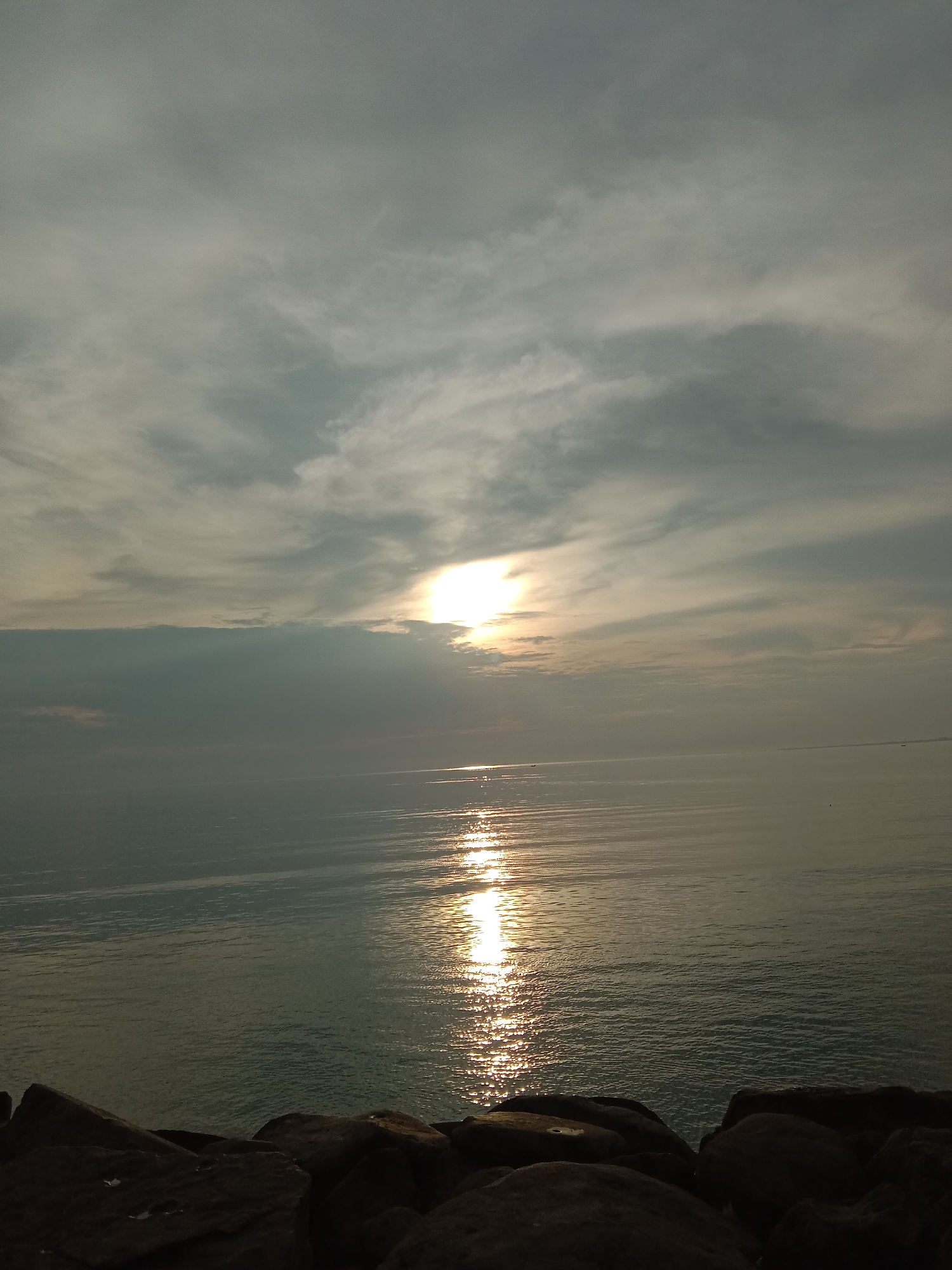 The image shows a seascape at dusk or dawn, likely taken in Lhokseumawe, Indonesia, as indicated by the prompt. The sun is partially obscured by a layer of clouds, creating a shimmering reflection on the water's surface. The sky is overcast with a mix of gray and white clouds. In the foreground, a rocky coastline is visible, with dark-colored stones. The water appears calm, with gentle ripples reflecting the sunlight. No people are visible in the frame.