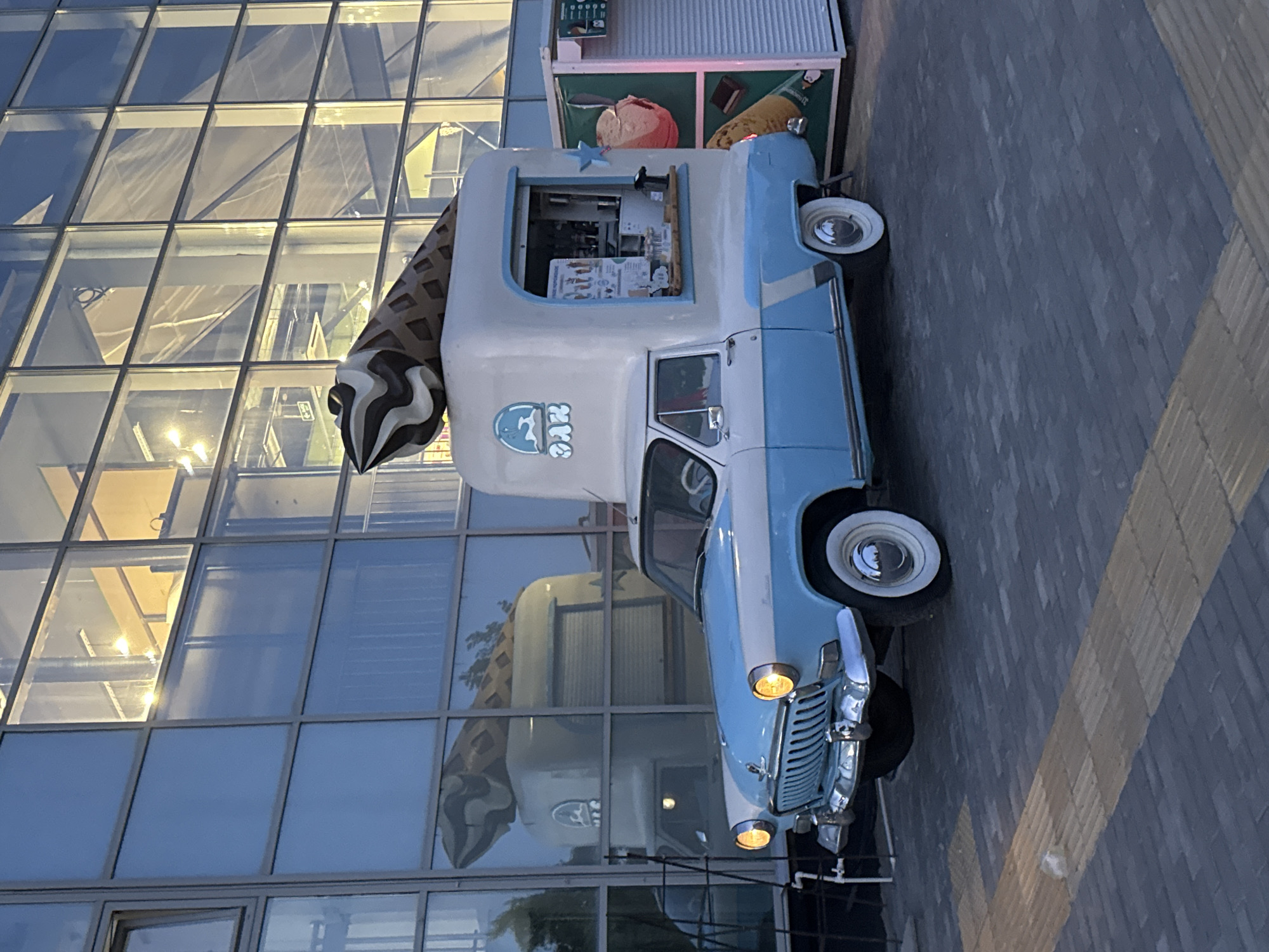 This media file captures an eye-level shot of a vintage ice cream truck parked outside a modern building with a glass facade. The truck is predominantly light blue and white, with a large, decorative ice cream cone emerging from its roof. The cone is brown and white striped, designed to resemble a classic ice cream cone. The truck itself appears to be a converted classic car, possibly a GAZ model given the logo on the side, which reads "ГАЗ" within a circular emblem.
The building's glass exterior reflects the truck and the surrounding environment, including the blue sky and some greenery, suggesting an outdoor setting. The reflections on the building are layered, showing the interior of the building with lights and structural elements, as well as the exterior scene. The truck has a serving window, indicating its function as an ice cream vendor. Inside the window, a menu with images of ice cream is visible.
The scene appears to be set during twilight or early evening, as indicated by the soft lighting and the dimming sky. The ground is paved with cobblestones, forming a walkway. There are no people visible in the frame. The overall mood is somewhat nostalgic, due to the vintage truck, contrasted with the sleek, contemporary architecture of the building. The location cues are minimal, but the presence of the GAZ logo, combined with the provided location context of Novosibirsk, Russia, helps to identify the origin of the vehicle.