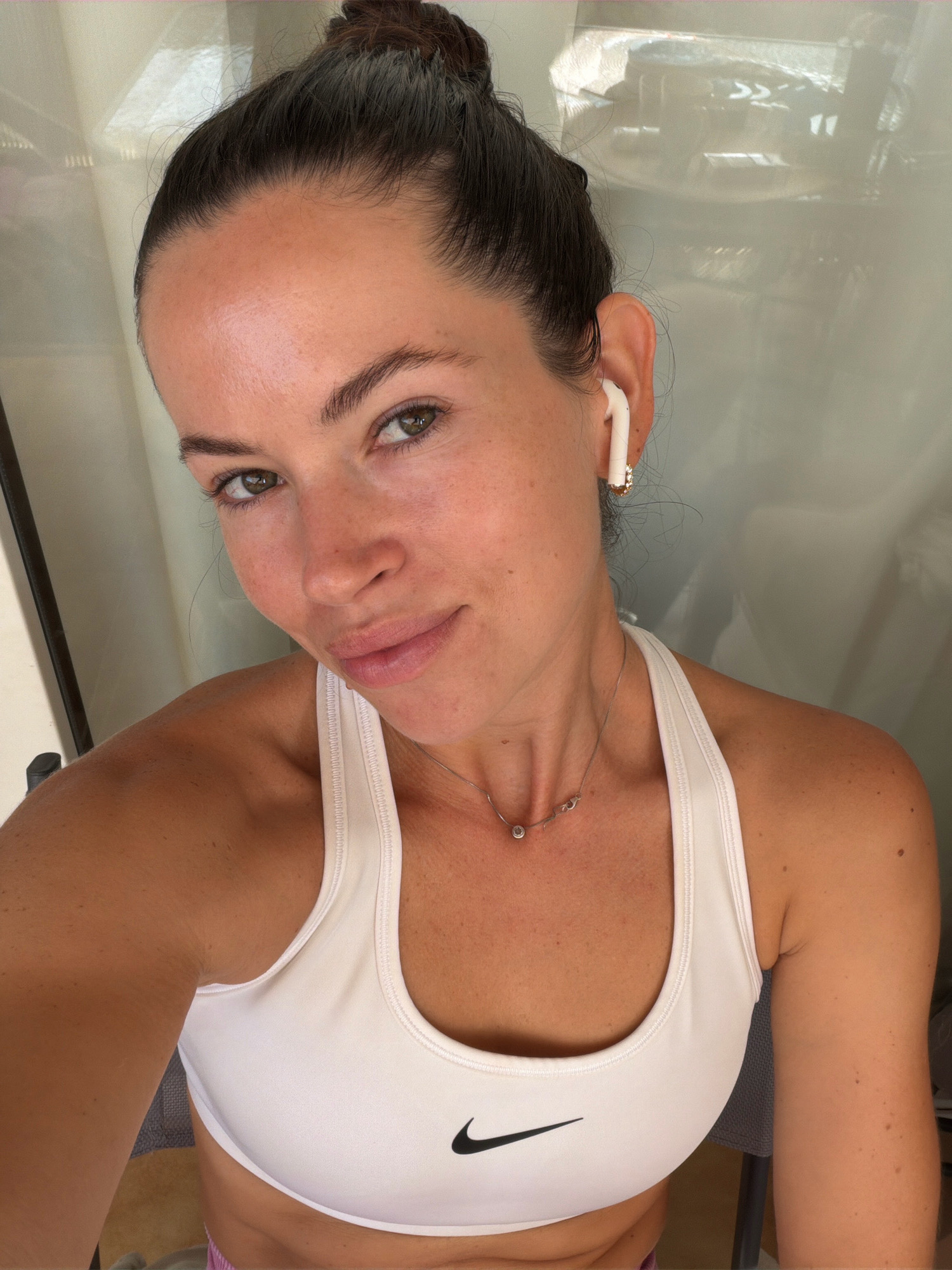 In Salou, Spain, a young woman with her hair in a bun and wearing white wireless earbuds poses for a selfie. She is sporting a white Nike sports bra and a delicate silver necklace. The backdrop appears to be a dimly lit indoor space with sheer curtains. The lighting suggests it's daytime, potentially late afternoon or early morning.