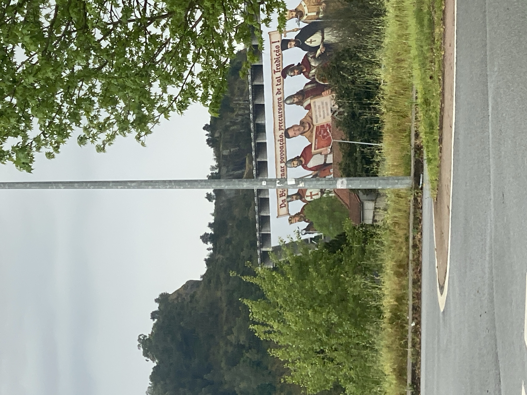 The image depicts a roadside scene in Braga, Portugal. A large mural is visible on a building or structure. The mural features a historical depiction of figures in period clothing, potentially related to the region's history or culture. A sign on the mural reads "Da B...nar povoação, precursora de tal Tradição!". The background includes a hillside covered in vegetation. A tree and a metal pole are in the foreground, slightly obscuring the view. A road is visible on the right side of the image. The sky appears overcast.