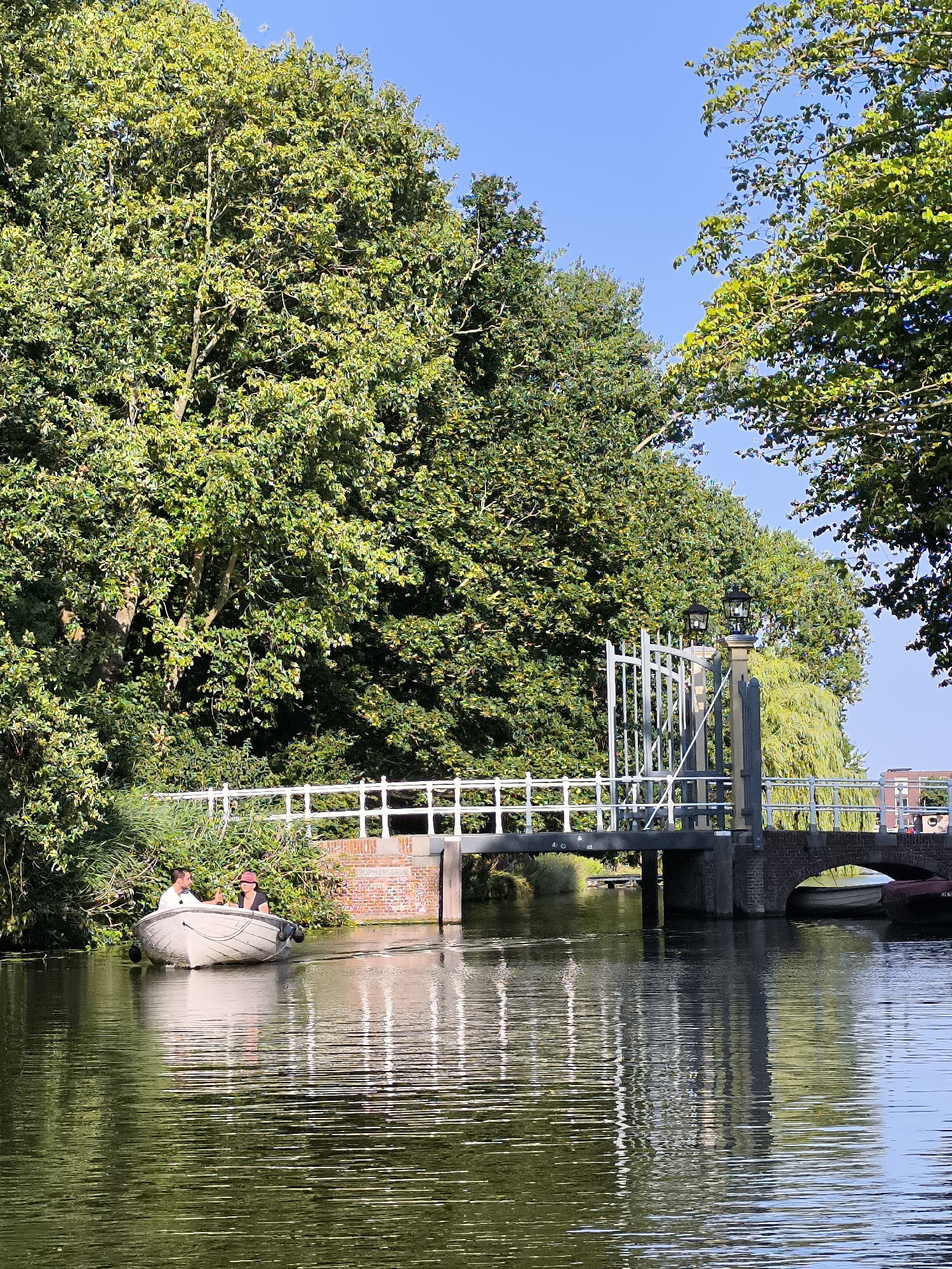 An outdoor, daylight, vertical, full shot captures a picturesque canal scene in Alkmaar, Netherlands. The water is calm, reflecting the surrounding lush green trees and a white bridge with ornate iron railings and lampposts. In the foreground, a small white boat carrying two individuals is moving from left to right. One person, wearing a white shirt, is steering, while the other, in a dark pink top and hat, appears to be speaking to them. The boat leaves a subtle wake on the water. The bridge spans the canal, with a brick structure supporting its walkway. Beyond the bridge, buildings and more trees are visible in the background under a clear blue sky. The overall impression is one of tranquility and a pleasant summer day.