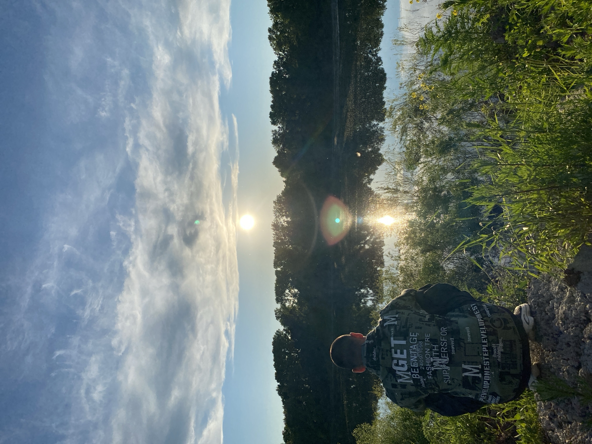 The image captures a serene outdoor landscape, likely at sunrise or sunset, by a body of water. A person is central to the foreground, viewed from behind and positioned on the grassy bank. They are crouching or sitting low, looking out over a calm river or lake. The individual wears a dark, camouflage-patterned jacket covered with various white text and numbers.
The bright sun, positioned on the left side of the frame, casts a strong glare and visible lens flares, with its reflection clearly visible on the water's surface. The far bank is lined with dense, dark green trees, their forms subtly mirrored in the calm water. The sky above is a clear light blue, accented by scattered, wispy white clouds. The immediate foreground features lush green grass and reeds, brightly illuminated by the warm, low-angle sunlight. The person's posture suggests a moment of peaceful observation or contemplation of the natural scenery.
Text visible on the jacket, as identified by OCR, includes phrases such as "MGETM", "BEENTAGE", "FASHION TRE WITH SUPPLIERSFOR", and "PLEVELUPONESTEPLEVELUPINEST". Other words like "debinti", "Mabini", "240971098", "sanh", and "Погон" are also present. The tranquil scene is set in Chaikovsky, Russia.