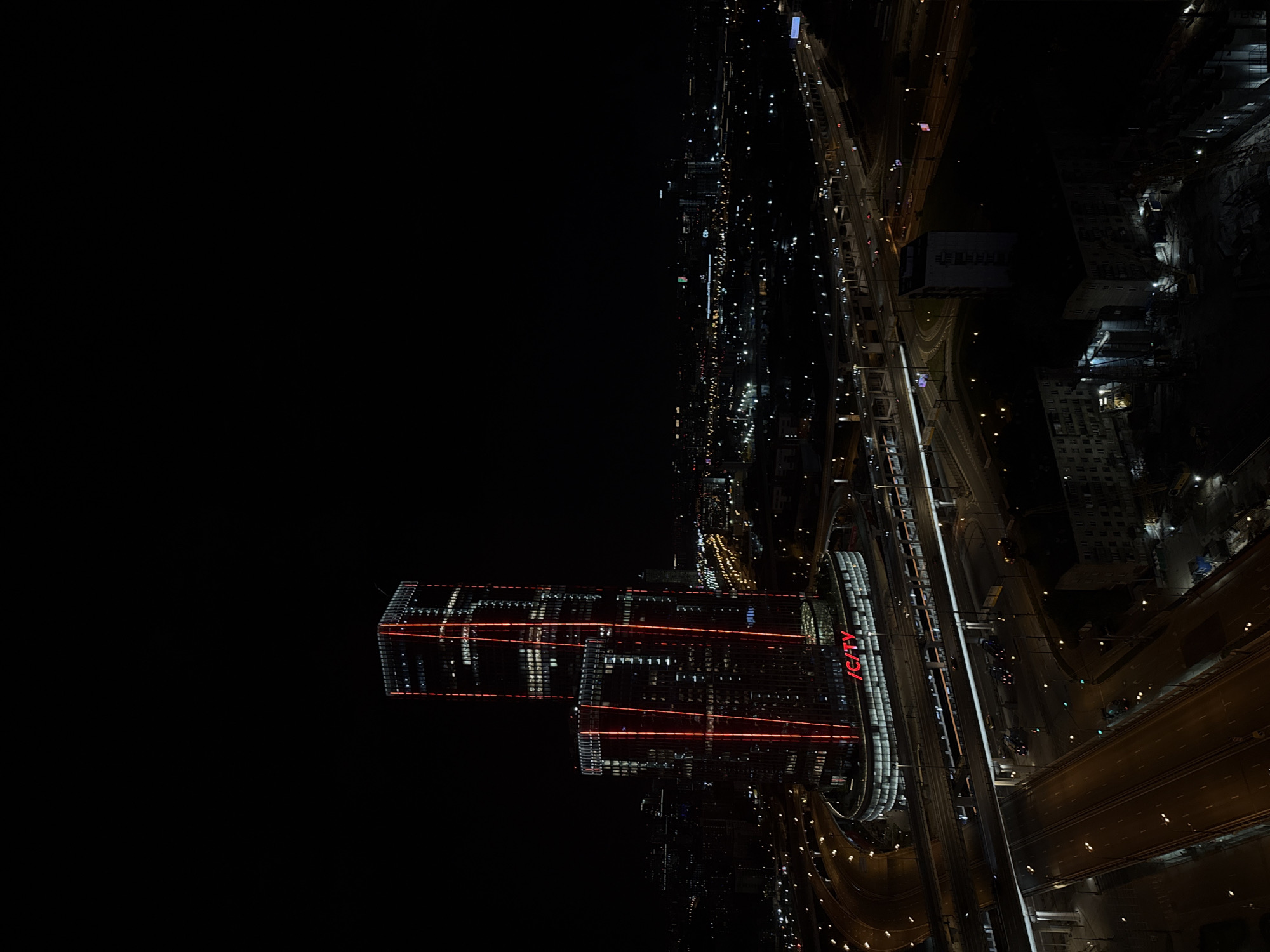 This is a high-angle or aerial nighttime photograph showcasing a vibrant urban landscape in Khimki, Russia. The scene is dominated by the stark contrast between the dark sky and the bright artificial lights of the city.
The primary subjects are two prominent, modern high-rise buildings located on the left side of the frame. These buildings are intricately illuminated with striking red horizontal and vertical light patterns, creating a distinctive architectural feature. On the facade of one of these buildings, the word "ICITY" is clearly visible in large, red, neon-like text.
To the right of the illuminated buildings, an extensive network of multi-lane highways, ramps, and overpasses winds through the city. These roads are brightly lit by numerous streetlights, and the presence of vehicle headlights and taillights, appearing as short light streaks, indicates active traffic flow. Other less distinct city buildings, some with illuminated windows, line the roads and extend into the background. A large, circular or curved structure, potentially an architectural element or a part of the road infrastructure, is also visible near the "ICITY" building.
The image captures the city at night, with the darkness emphasizing the glow of artificial lighting from buildings, streets, and vehicles. The weather appears clear, with no visible atmospheric conditions. No individual people are discernible, but the bustling lights and traffic suggest a lively urban environment.