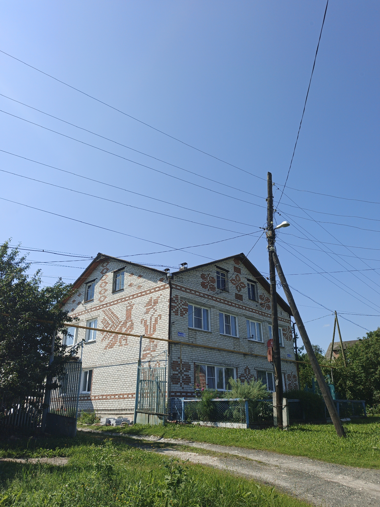 In Medvedyovka, Russia, a three-story brick building with decorative orange and white patterns on its facade stands under a clear blue sky. The building has numerous windows on each floor. In front of the building, a metal gate is open, leading to a dirt path. Green grass and trees surround the area, with a dense tree line visible to the right. A utility pole with electrical wires and a street lamp stands prominently in the foreground to the right of the building, leaning slightly. The scene appears to be a residential area on a bright, sunny day.