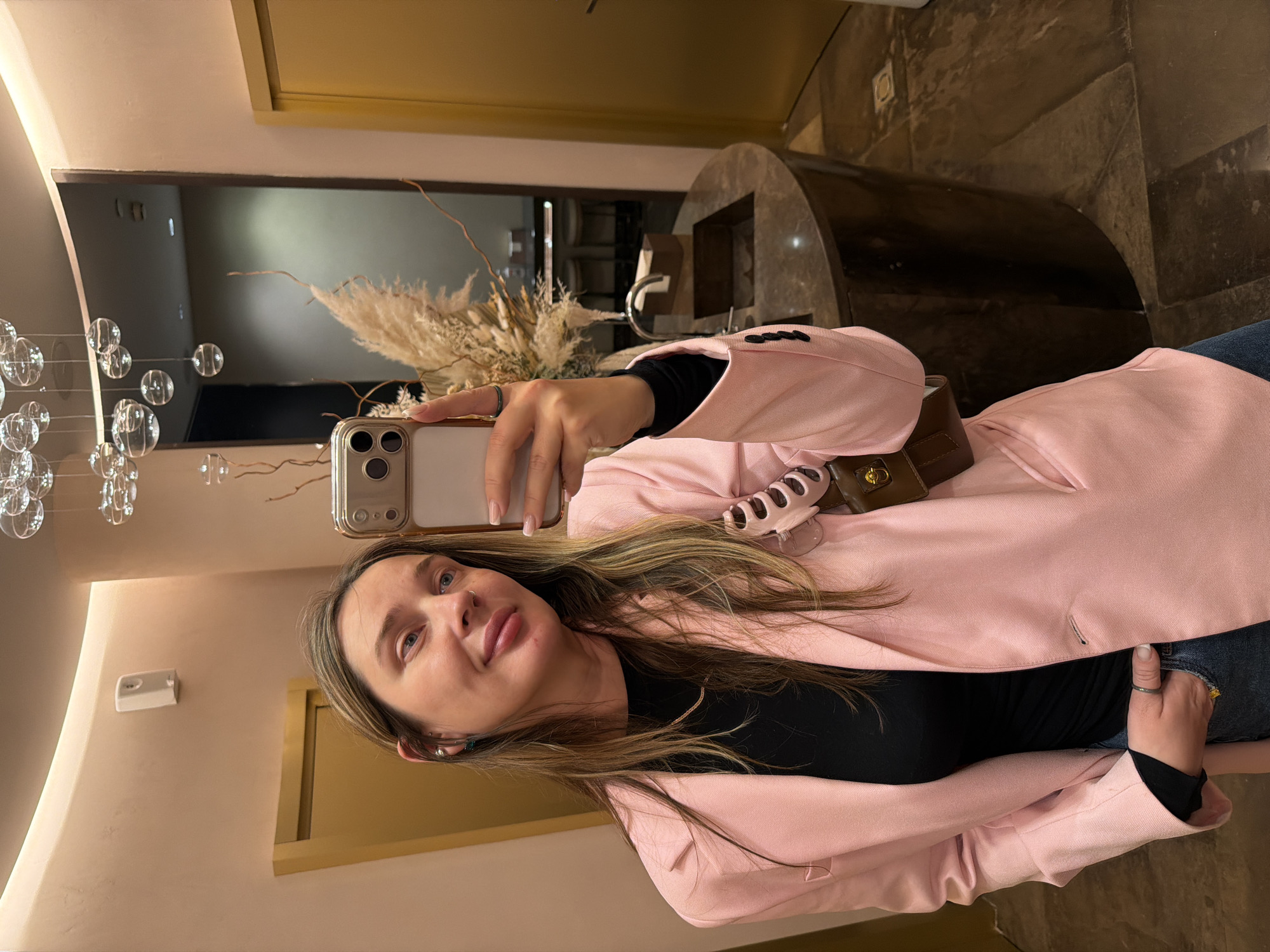 A woman is taking a selfie in a restroom. She is wearing a pink blazer over a black top and jeans. She has long, blonde hair and is wearing earrings and a ring. In the background, there is a sink, a decorative plant, and a door. The overall impression is one of modern elegance, possibly in a restaurant or boutique hotel. The image does not provide any specific clues that would indicate the location within Saint Petersburg, Russia.