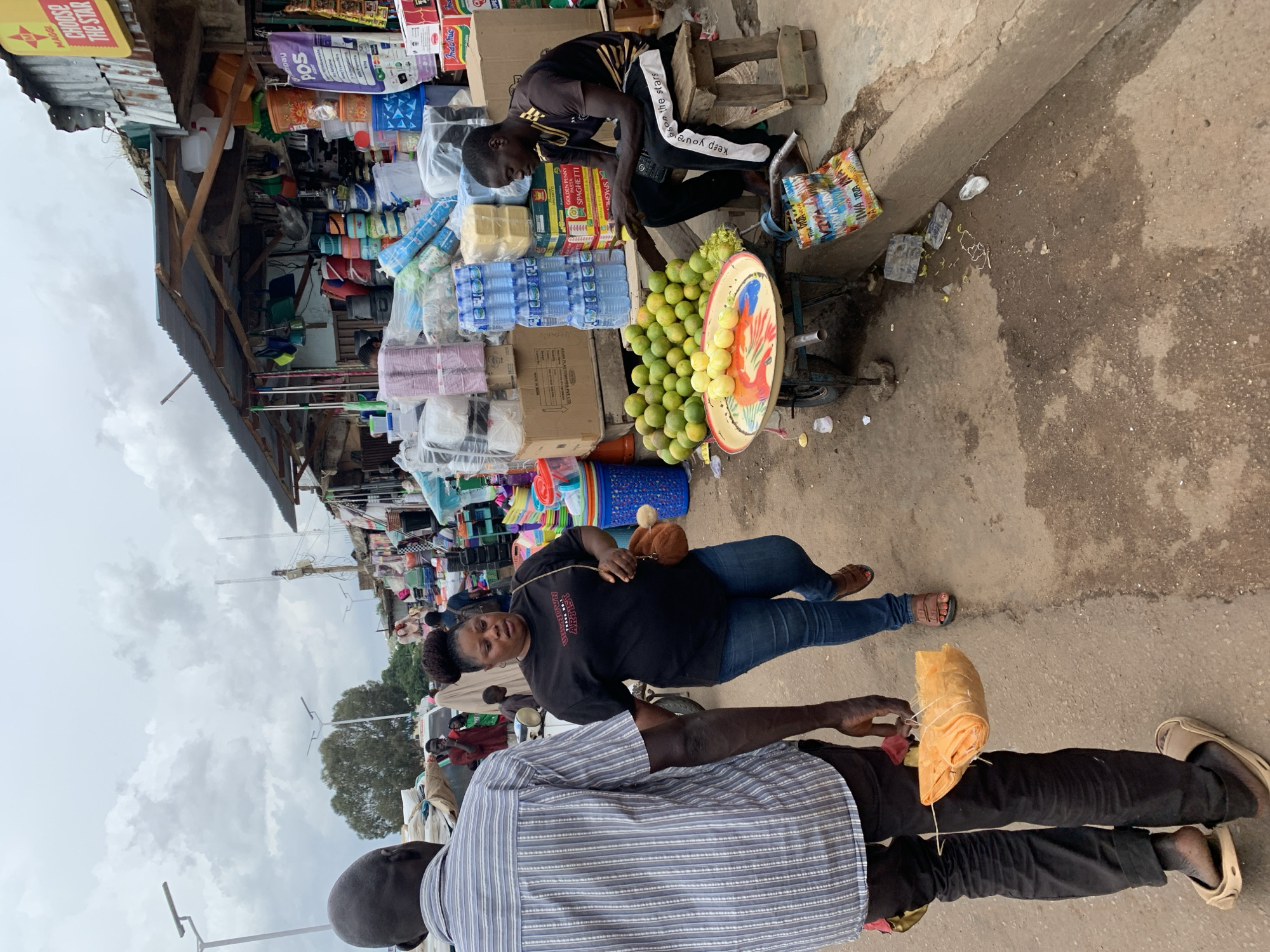 The image depicts a busy street market scene in Jos, Nigeria, under a cloudy sky. In the foreground, a man wearing a striped shirt and dark pants is walking away from the camera, carrying a bundle wrapped in orange material tied with string. To his right, a woman in a black t-shirt and blue jeans is walking towards the right side of the frame. She has her hair styled in a bun and is carrying a small brown bag on her shoulder.
The background reveals a row of market stalls. One stall on the right is covered with a corrugated metal roof supported by wooden beams. It displays various goods, including stacks of bottled water, detergent, and packaged food items like spaghetti. A man is seated on a wooden stool in front of this stall, looking at something in his hand. In front of him, a cart is laden with a large pile of green citrus fruits, possibly limes or small oranges, and a smaller pile of yellow, round fruits, perhaps lemons. A colorful patterned bag is placed on the ground near the cart. Further down the street, other market stalls and people are visible, indicating a bustling commercial area. The ground appears to be a dirt or asphalt road with some puddles and debris. Streetlights are visible in the background, suggesting it is an urban setting.