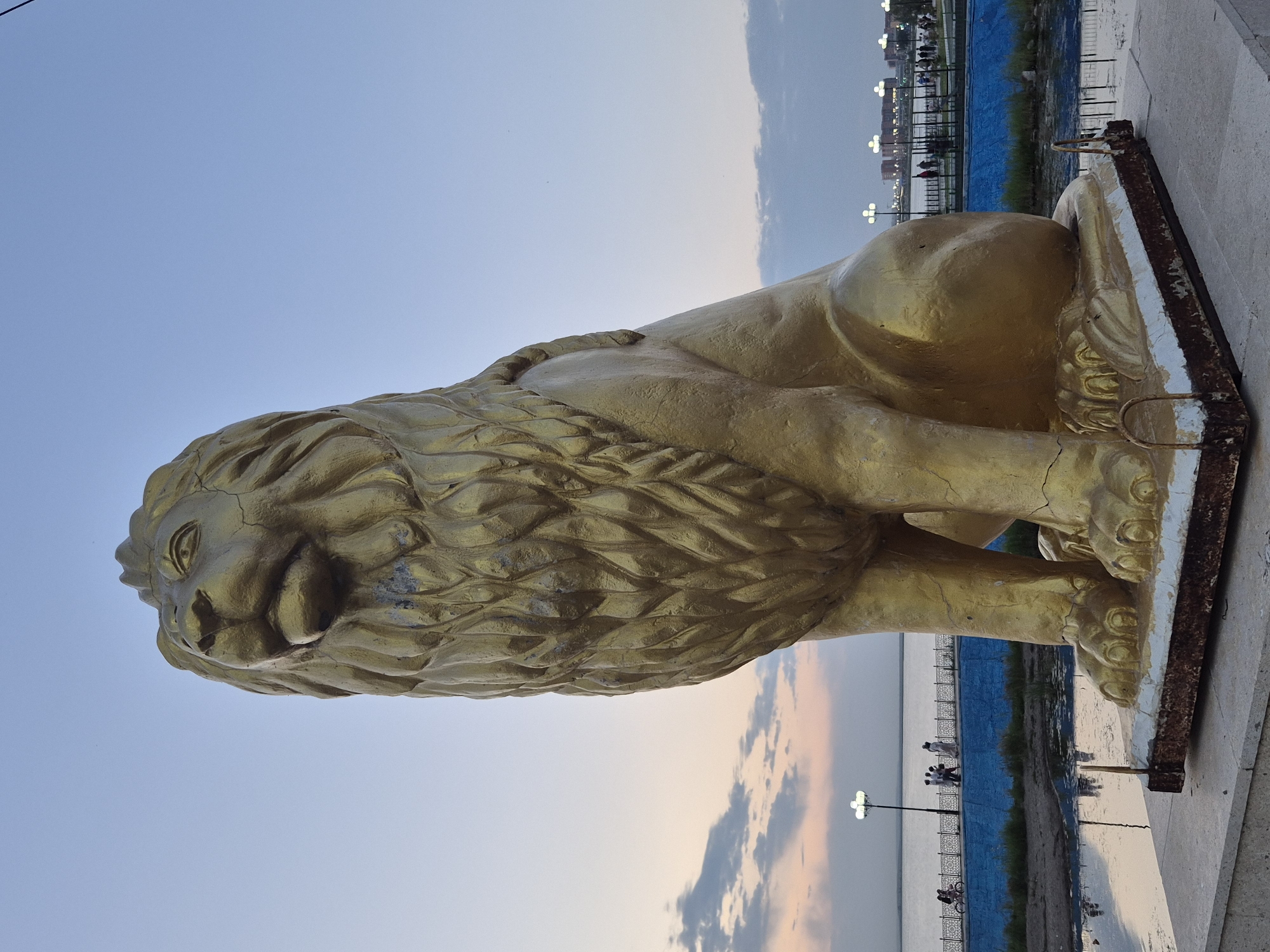 A golden lion statue sits prominently in the foreground, facing left, with its head turned upwards. The statue is weathered, showing cracks and discoloration, particularly on its mane and paws. Behind the statue, a body of water stretches towards the horizon. A walkway with a blue barrier runs along the edge of the water, illuminated by streetlights. Several people are visible on the walkway, some walking and one riding a bicycle. In the distance, buildings and more streetlights suggest an urban environment. The sky is a blend of soft blues and pinks, indicating either dawn or dusk. The overall scene suggests a public park or promenade by the water. The statue is a prominent landmark in Kokshetau, Kazakhstan.