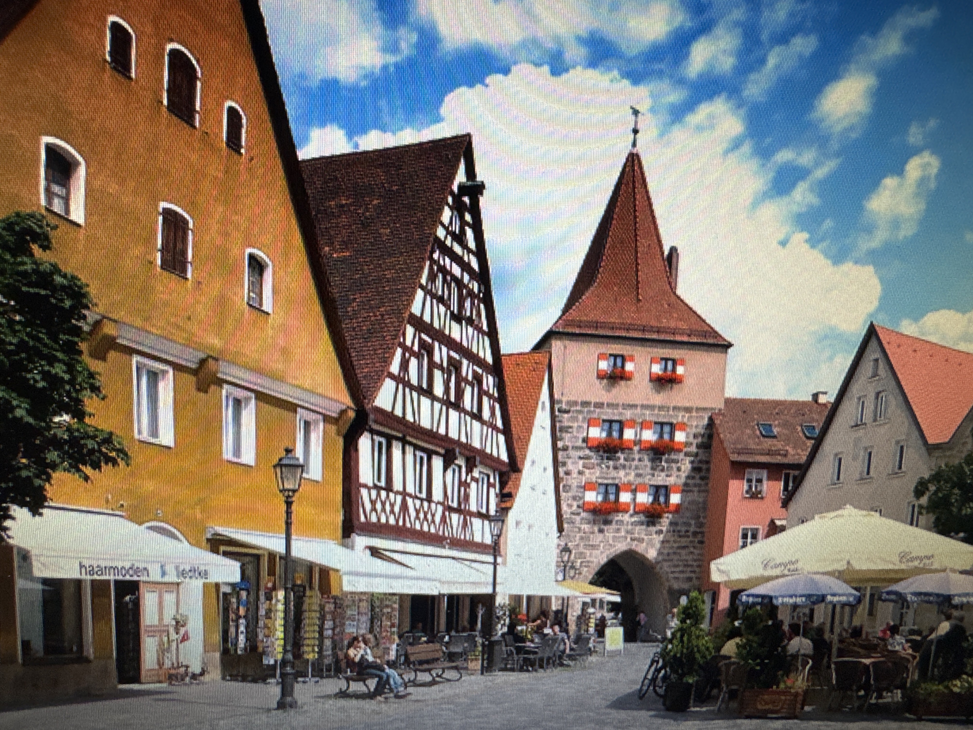 The image shows a street scene in Ellwangen, Germany, specifically the Schulkellerberg gate and surrounding buildings. The gate, a prominent stone structure with red-and-white window shutters, is centrally located. To its left, a half-timbered building with a steeply pitched, tile-covered roof features decorative wooden beams. Further left, a bright yellow building with multiple arched windows and a flat roof is visible.
In the foreground, several shops with awnings line the street. One awning displays the text "haarmoden Liedtke". A traditional street lamp stands in front of the shops. Benches are placed along the walkway, with a few people seated on them. Outdoor cafe seating with umbrellas is set up in front of other establishments, with patrons seated at tables. A bicycle is parked near the cafe area. The sky is blue with scattered white clouds. The overall impression is of a charming, historic European town square.