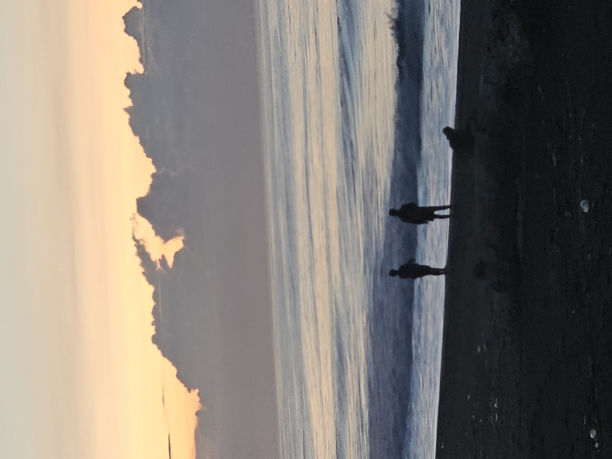 The image shows a coastal scene in Denpasar, Indonesia, possibly during sunset. Three silhouetted figures stand on a dark sandy beach near the waterline. Waves break on the shore, and the sky transitions from a pale orange-yellow near the horizon to a darker gray towards the top of the frame. Clouds are visible near the sunset.