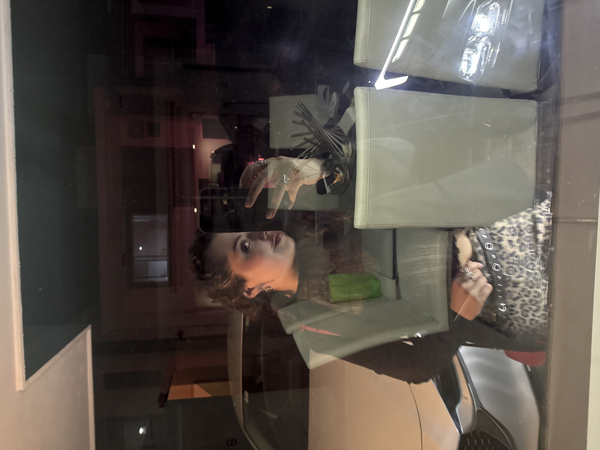 A woman is reflected in a glass window. She is holding a smartphone with her right hand, her fingers slightly splayed. She is wearing rings on her fingers. Behind her is a room with a white chair and a shelf with decorative objects. The reflection in the window shows buildings across the street. The location in Santa Clara Ocoyucan, Mexico cannot be confirmed from the image.
