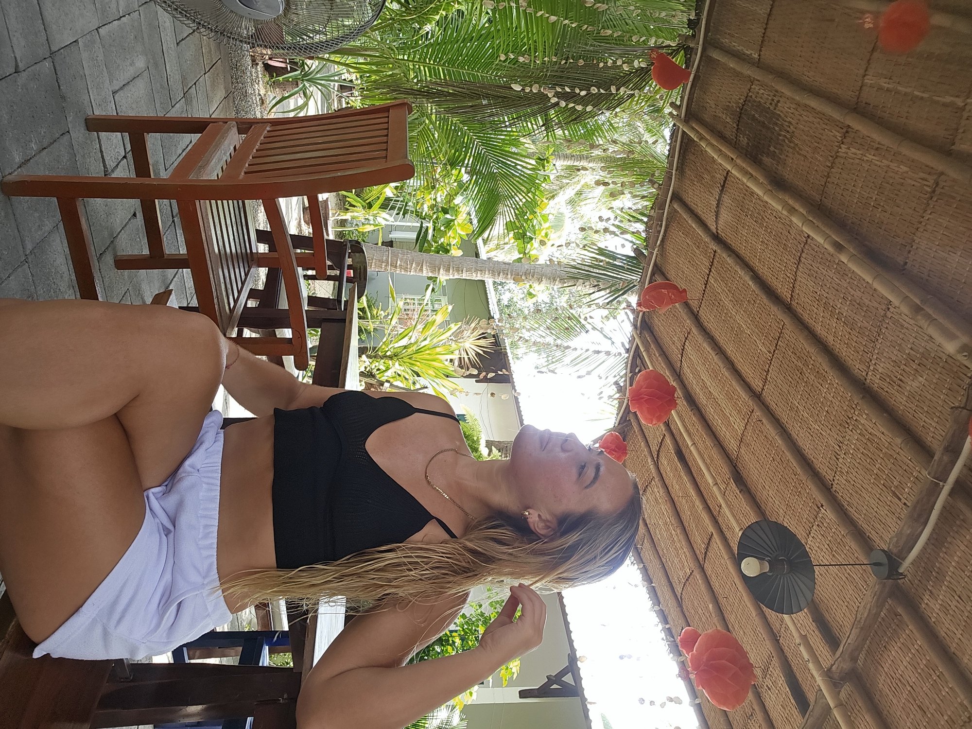 A woman with blonde, wet hair is seated in an outdoor cafe in Phu Thuy Ward, Vietnam. She is wearing a black crop top and white shorts. The cafe has a thatched roof and is surrounded by lush greenery, including palm trees and other tropical plants. Red paper lanterns hang from the ceiling, and a black pendant light is visible. Wooden chairs and tables are present. The flooring is paved with grey rectangular tiles. A fan is positioned to the left of the frame. The woman is looking to her right, with her hand touching her hair. The overall atmosphere suggests a relaxed, tropical setting.