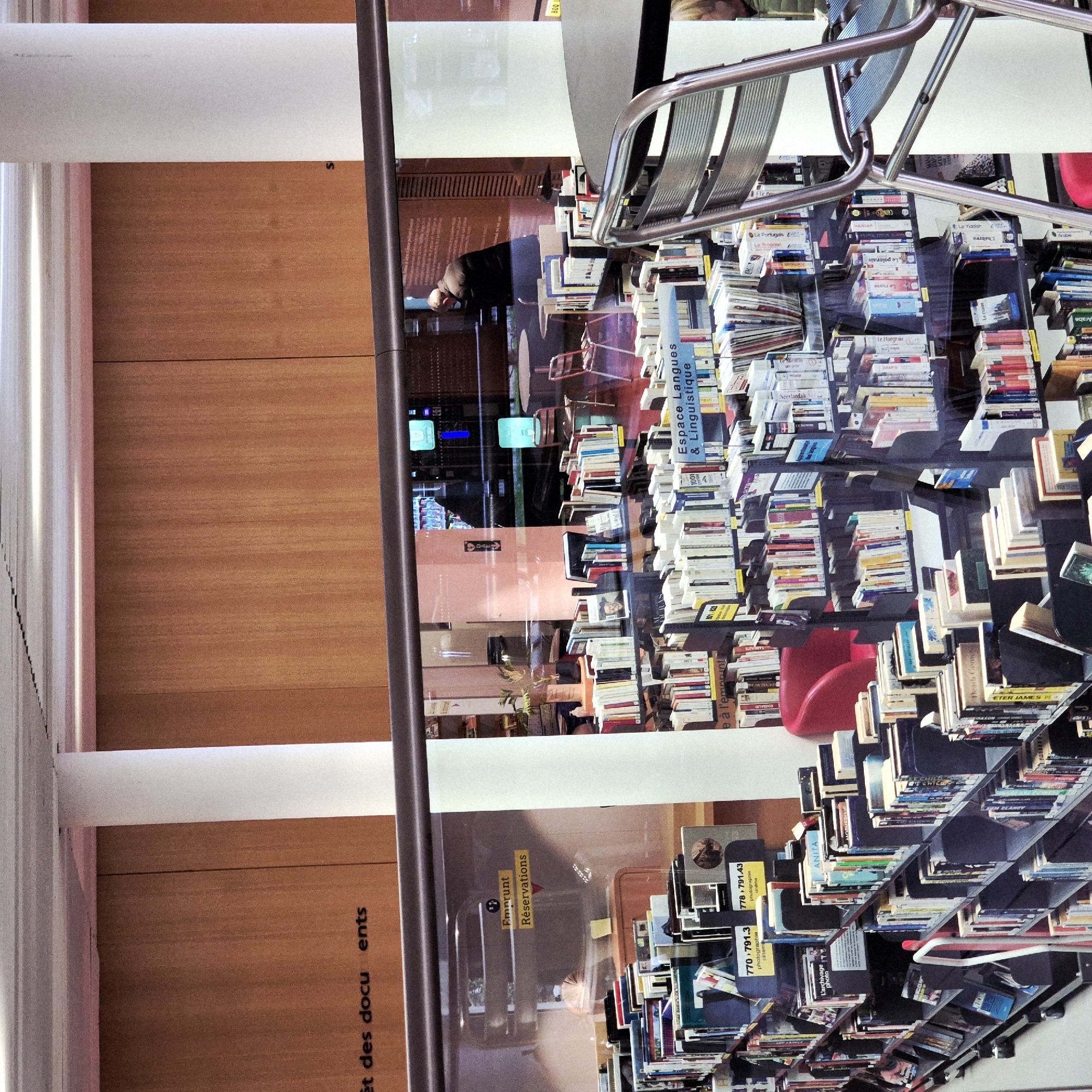 This image depicts a reflection of a library setting, likely in Strasbourg, France, based on the French text visible. The scene features stacks of books labeled in various languages ("Espace Langues & Linguistique"), a metal chair, and a red chair near the bookshelves. Reflected in the glass is a person standing in the library's interior, along with interior signs indicating "Emprunt Réservations" (Borrowing Reservations). The reflected environment suggests a modern library space, with visible bookshelves, seating, and signage related to borrowing services. The scene captures the interaction of people and objects within a library environment focused on language and linguistics. The books cover a variety of topics, including photography and cinema, as indicated by the call numbers.