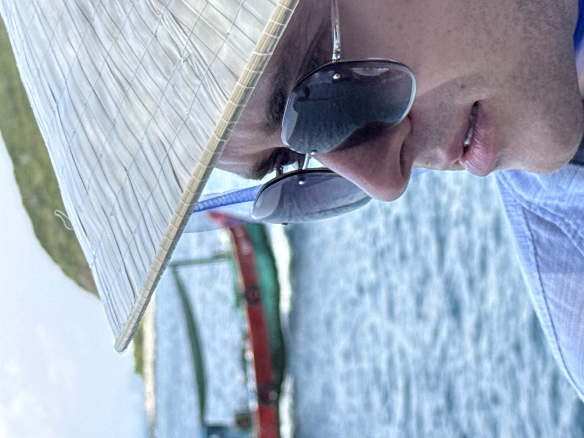 The image shows a person wearing a traditional Vietnamese conical hat and sunglasses, with their face in profile. The person is on a boat, as suggested by the water and a section of the boat's railing visible in the background. The setting appears to be outdoors during the daytime. The location is Nha Trang Ward, Vietnam.