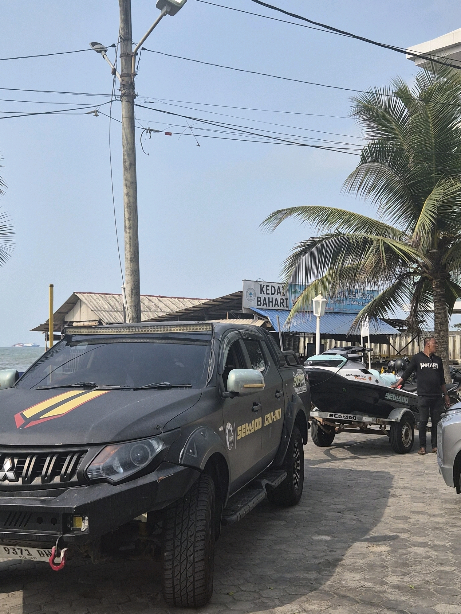 In Balikpapan, Indonesia, a black Mitsubishi Triton pickup truck is parked on a paved area near the sea. The truck features a custom black bumper with a red tow hook, a light bar mounted above the windshield, and yellow and red racing stripes on the hood. Next to the truck, a jet ski on a trailer is visible. A man wearing a black t-shirt with the word "NOTARY" and black pants stands near the trailer, looking to his right. Behind the vehicles, a building with a sign that reads "KEDAI BAHARI" (Marine Cafe) is situated, with palm trees and power lines in the background. The ocean is visible in the distance, with two boats on the water. The overall scene depicts a coastal area with recreational vehicles and a local establishment.