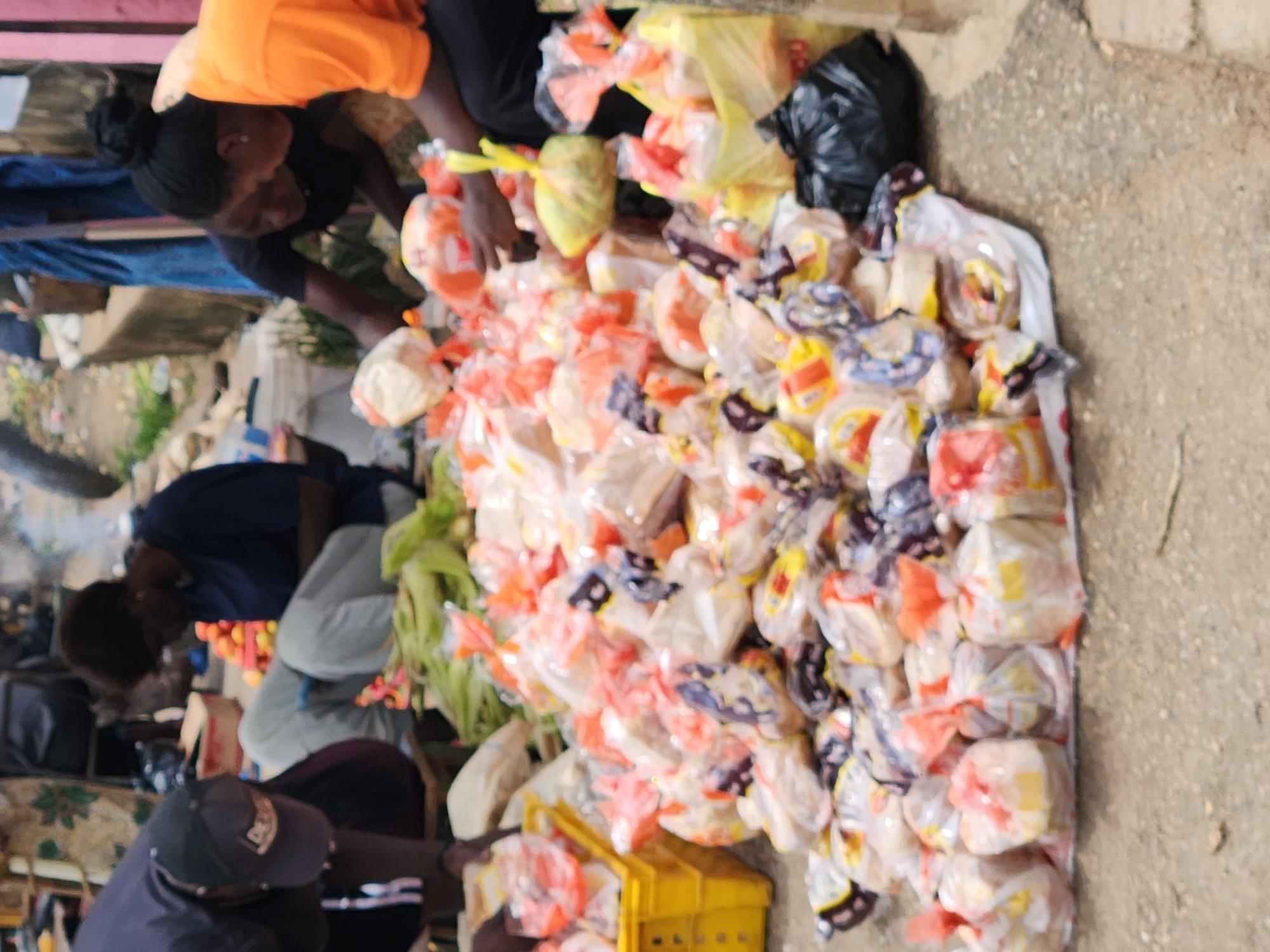 The image depicts a bustling market scene in Kabong, Nigeria. In the foreground, a large pile of individually wrapped loaves of bread is spread out on the ground, contained within a white plastic sheet. The bread is packaged in clear plastic with some featuring red and orange ties or labels. A yellow plastic crate is visible to the left, also containing bread. Several individuals are present in the scene. On the left, a person wearing a dark blue shirt and a baseball cap is bending down, potentially tending to the goods. In the center, a woman in a dark blue t-shirt is seated, looking down at her phone. To her right, another woman in an orange t-shirt is also seated and appears to be interacting with the bread. In the background, there are hints of other market stalls, produce, and possibly more people, contributing to a sense of vibrant commercial activity. The ground is paved with concrete or asphalt.