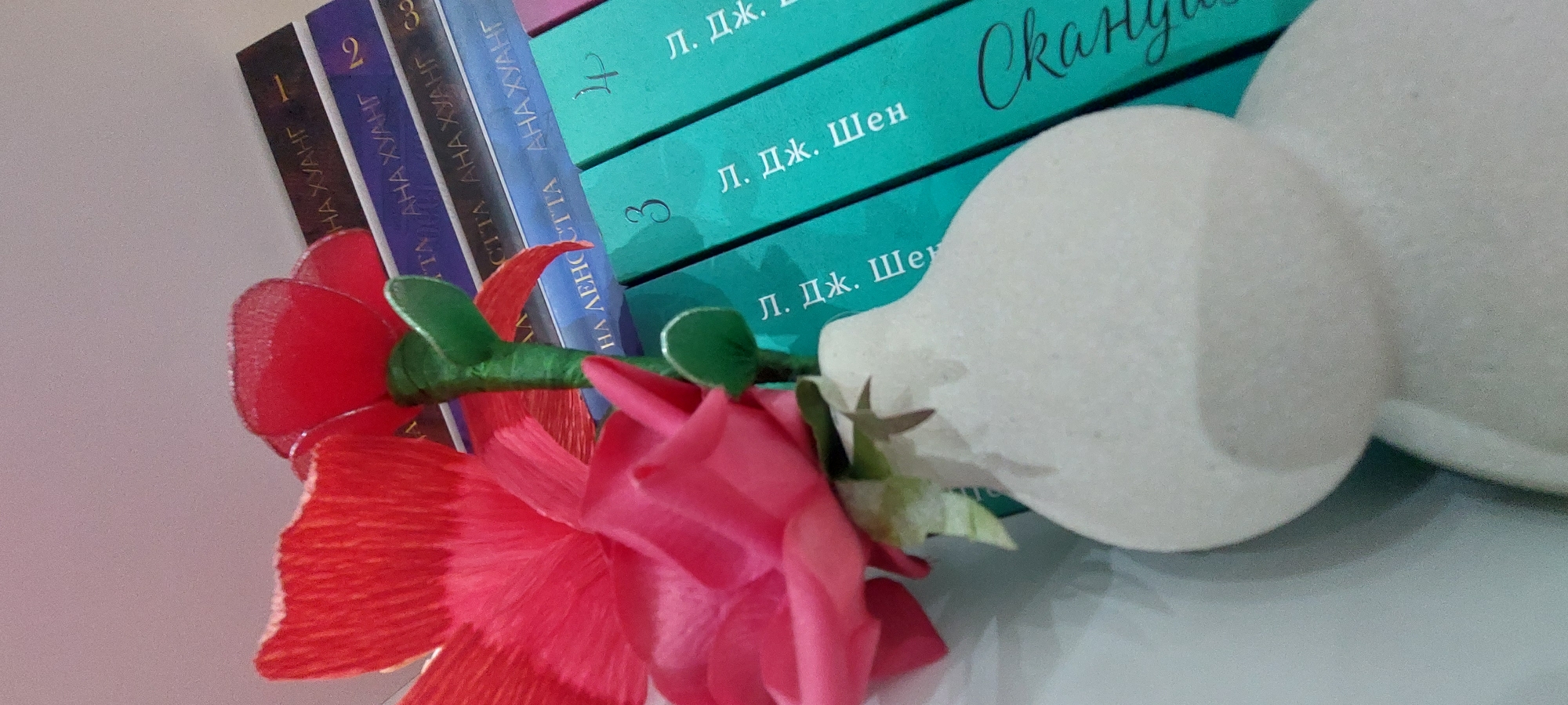 The scene is depicted in the city of Yagodovo, Bulgaria.
The image presents a still life composition featuring a white vase holding artificial flowers, positioned in the foreground, with a stack of books arranged diagonally in the background.
The vase, occupying the lower-left portion of the frame, is an off-white, matte-textured object with an irregular, organic, bulbous shape that tapers towards a narrower neck at the top.
Two primary artificial flowers are visible in the vase. The prominent flower in the immediate foreground is a large, ruffled bloom of vibrant pink or magenta, constructed from a fabric or crepe paper-like material. Behind it, and slightly to the right, is a smaller, glossy red flower with distinct, somewhat heart-shaped petals, appearing to be made from plastic or a similar rigid material. Both flowers have green stems, wrapped in fabric, with small, pointed green leaf-like elements.
Behind the vase and flowers, a stack of six books is visible, arranged horizontally with their spines facing the viewer. The top three books have dark grey, dark blue, and light blue spines, respectively. Visible Cyrillic text on these spines includes "АНА ХУАНГ" (ANA KHUANG) with numerical indicators "1", "2", and a partial "3" on the light blue book. The dark blue spine also shows "НА ДЕНЮЭТТА" (NA DENYUETTA). The bottom three books have teal-colored spines and display "Л. ДЖ. Шен" (L. DZH. SHEN) along with numerical indicators "3" and "4". The rightmost visible teal spine also features the cursive Cyrillic word "Скандал" (Scandal).
The objects rest on an unidentifiable flat, light-colored surface. The background consists of a plain, uniformly light-colored wall or vertical surface. The lighting is soft and even, originating from an unseen source, and casts minimal shadows.