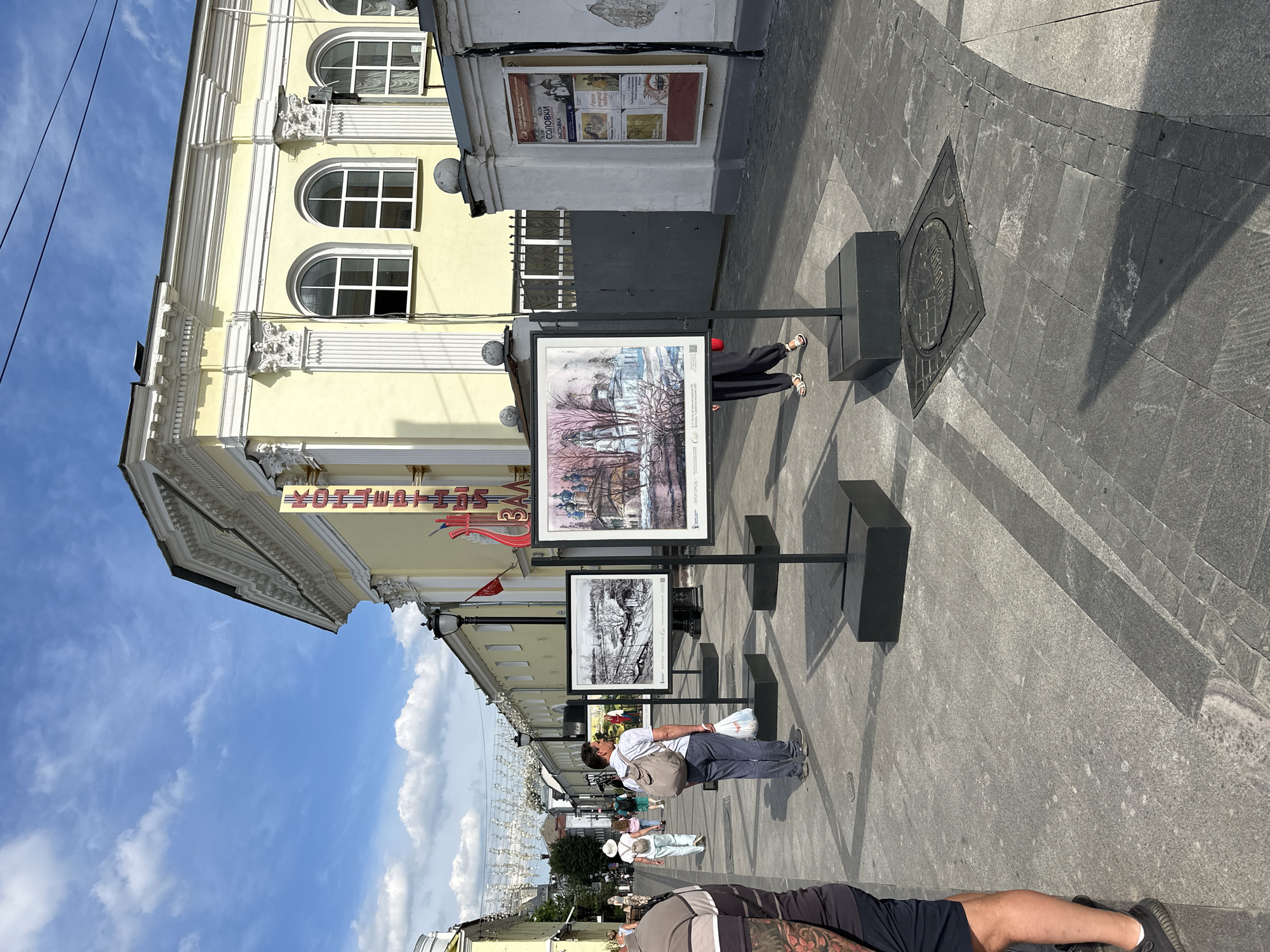 An outdoor art exhibition is set up on a paved walkway in front of a light yellow building. Several framed artworks are displayed on black stands. In the foreground, a person is visible from the waist down, wearing a dark t-shirt with a design and dark shorts. To the left, a woman in a white hat and a light-colored outfit walks away from the camera, carrying a white bag and wearing sandals. Behind her, another person in a gray backpack and dark pants is looking towards the artworks. Further down the walkway, more people are walking and the street is lined with buildings and lampposts. Overhead, decorative string lights are suspended across the walkway. The sky is blue with scattered white clouds. The building in the background has arched windows and decorative architectural elements. A poster is affixed to the wall of a white building on the right side of the image. The location is Putyatino (Semivragovskaya S/A), Russia.