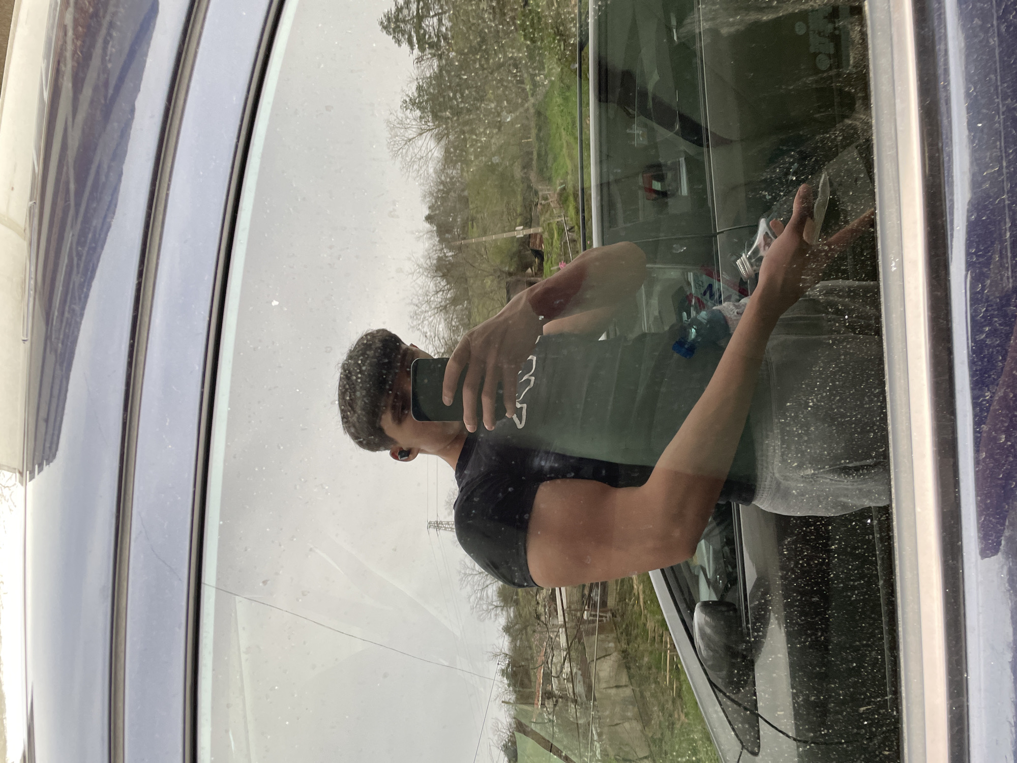 The image, oriented sideways, shows the reflection of a person taking a selfie in a dirty car window. The person, appearing male, is seen from the chest up, wearing a dark-colored t-shirt with some white, mirrored text or design (possibly starting with 'W') on it. They are holding a dark smartphone to capture their reflection, with an earbud visible in their right ear.
The car's dark blue or purple frame is visible on the left and right edges of the window. The window itself is heavily smudged and covered in numerous water spots, dust, and fine scratches, suggesting either recent rain or a dusty, overcast environment.
The reflected background reveals an outdoor setting with a natural landscape: bare trees, green grass, and some distant structures or utility poles under a bright, overcast sky. Hints of the car's interior, including a light-colored object, are faintly visible through the reflection. The scene is situated in Zlatograd, Bulgaria, under dull, diffused lighting conditions.
