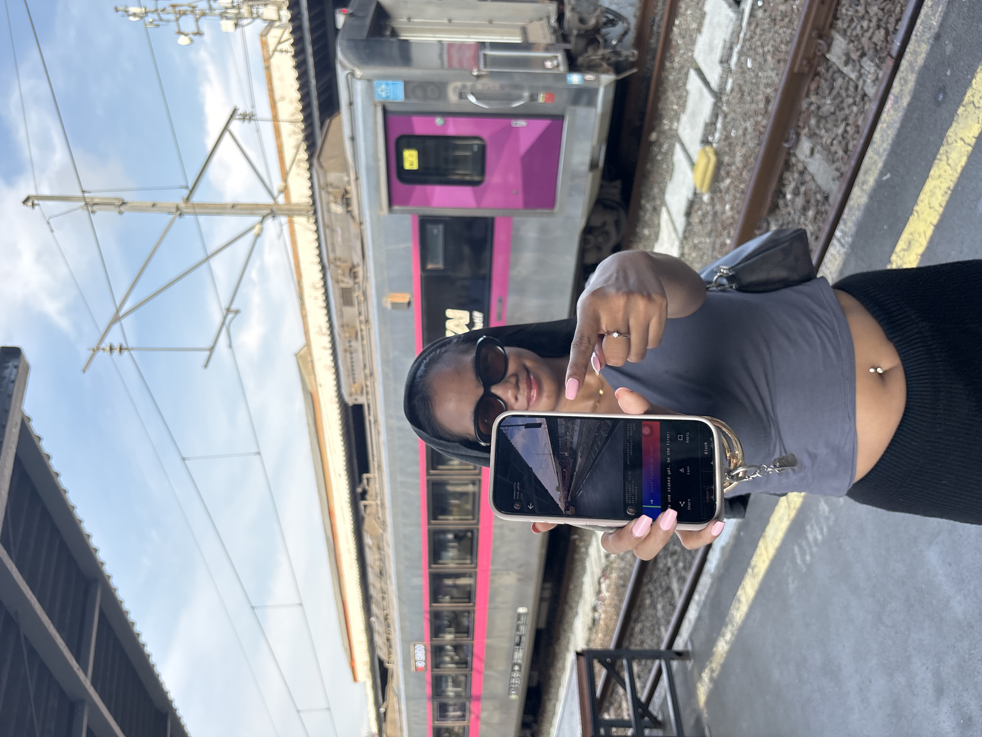 The image depicts a woman standing on a train platform, holding up her smartphone to take a picture of the train behind her. She is wearing sunglasses, a gray tank top, and black bottoms. A small black purse hangs over her shoulder. She has a ring on her finger and a navel piercing.
The train behind her is silver with a pink stripe and pink door. There is a set of tracks and gravel on the ground. Above the train and platform are overhead electrical lines and structures. The sky is visible in the upper left corner, partially obscured by the platform roof structure.
The smartphone screen shows what appears to be a photo editing or social media interface, with elements like a share button, save button, and user options.
Based on the visible train elements, this does not seem to be located in West Jakarta, Indonesia as the city doesn't utilize trains of this style.