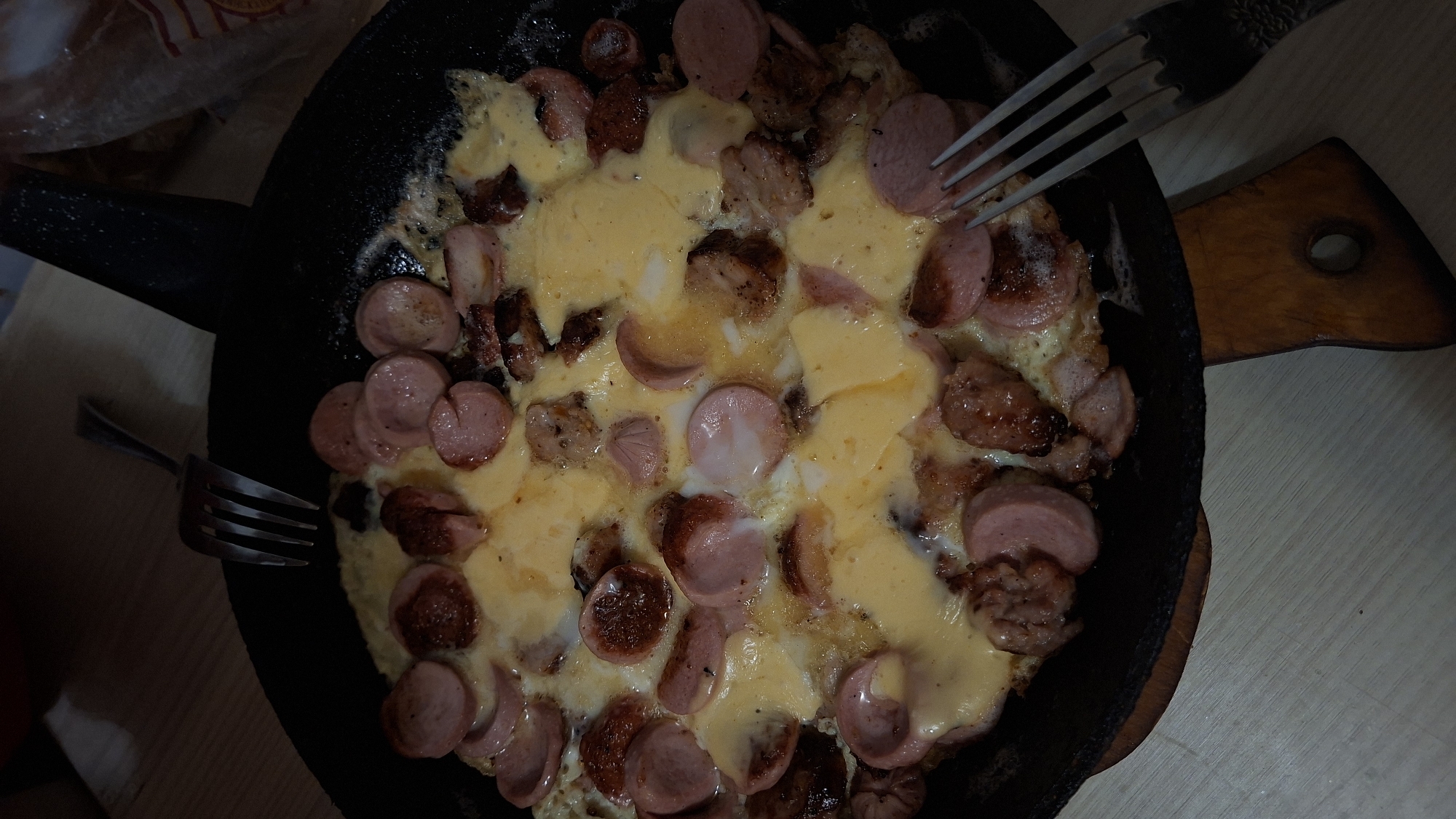 The image shows a close-up, overhead view of a skillet filled with a cooked dish, likely an omelet or scrambled eggs mixed with sliced sausages and melted cheese. The food is dark and appears to be in a domestic setting, possibly a kitchen or dining area. Two forks are visible, one on the left side of the skillet and another on the right, suggesting the meal is either being prepared or has been served. The lighting is dim, casting shadows and creating a somewhat muted color palette. The background is out of focus, but appears to be a table surface. There are no people visible in the image. The dish itself is a mixture of textures and colors, with the pinkish hue of the sausages, the yellow of the melted cheese, and the brown of some browned bits of meat or sausage. The overall impression is of a hearty, home-cooked meal. The mention of "Забалоцкі сельскі Савет, Belarus" in the prompt implies a rural setting in Belarus, which aligns with the rustic appearance of the food and the potential domestic environment.