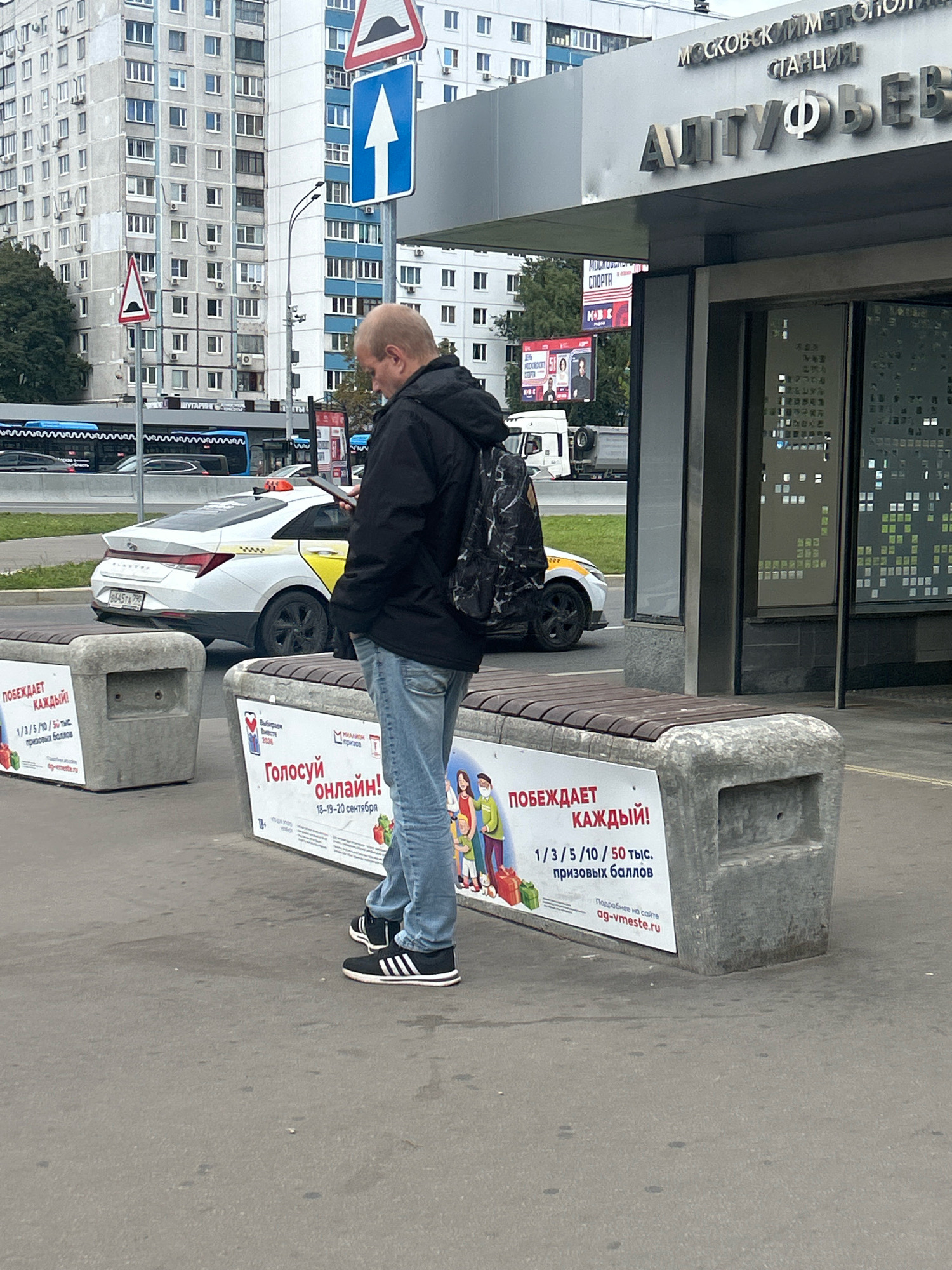 The image shows a man standing on a sidewalk in Moscow, Russia, near a bus stop and a metro station. The man is wearing a black jacket, blue jeans, and black and white sneakers. He is looking at his phone and holding a black backpack. In the background, there are several apartment buildings, a bus, and a white taxi. There are also two concrete benches with advertisements on them. One advertisement says "ГОЛОСУЙ ОНЛАЙН!" which translates to "VOTE ONLINE!". Another advertisement says "ПОБЕЖДАЕТ КАЖДЫЙ!" which translates to "EVERYONE WINS!". The metro station sign says "МОСКОВСКИЙ МЕТРОПОЛИТЕН СТАНЦИЯ АЛТУФЬЕВО" which translates to "MOSCOW METRO STATION ALTUFIEVO". The overall scene suggests an urban environment on a cloudy day.