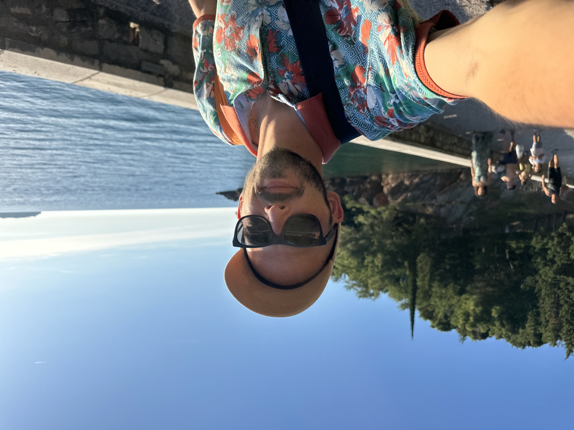 A man, wearing sunglasses and a cap, takes a selfie outdoors. He is wearing a floral-patterned shirt and has a beard. Behind him, the blue sea stretches out, with a stone wall and lush green trees visible. In the background, a few people are walking along a path. The scene appears to be located in Sveti Stefan, Montenegro, a coastal town known for its picturesque views.