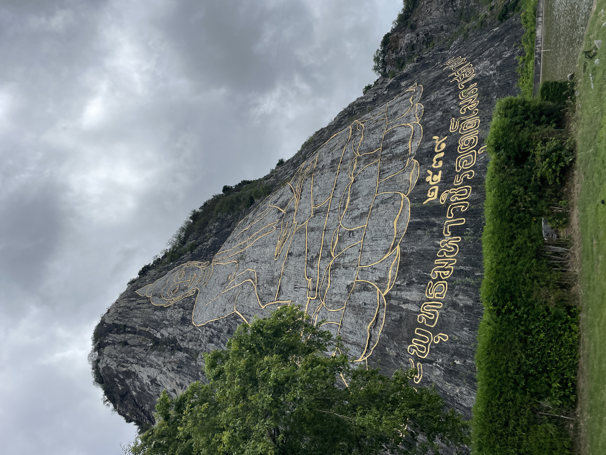 An enormous golden outline of a seated Buddha figure is inscribed on the side of a large, grey, rocky mountain. The Buddha image is very large, taking up a significant portion of the mountainside. Below the Buddha, there is text in a golden script. The mountain is partially covered with some green trees and vegetation on its upper slopes. In the foreground, there is a neatly trimmed green hedge, and beyond that, a patch of grass. To the right of the hedge and grass, there is a body of water. The sky above is overcast with grey clouds. This is the Khao Chi Chan, or Buddha Mountain, located in Na Chom Thian, Thailand.