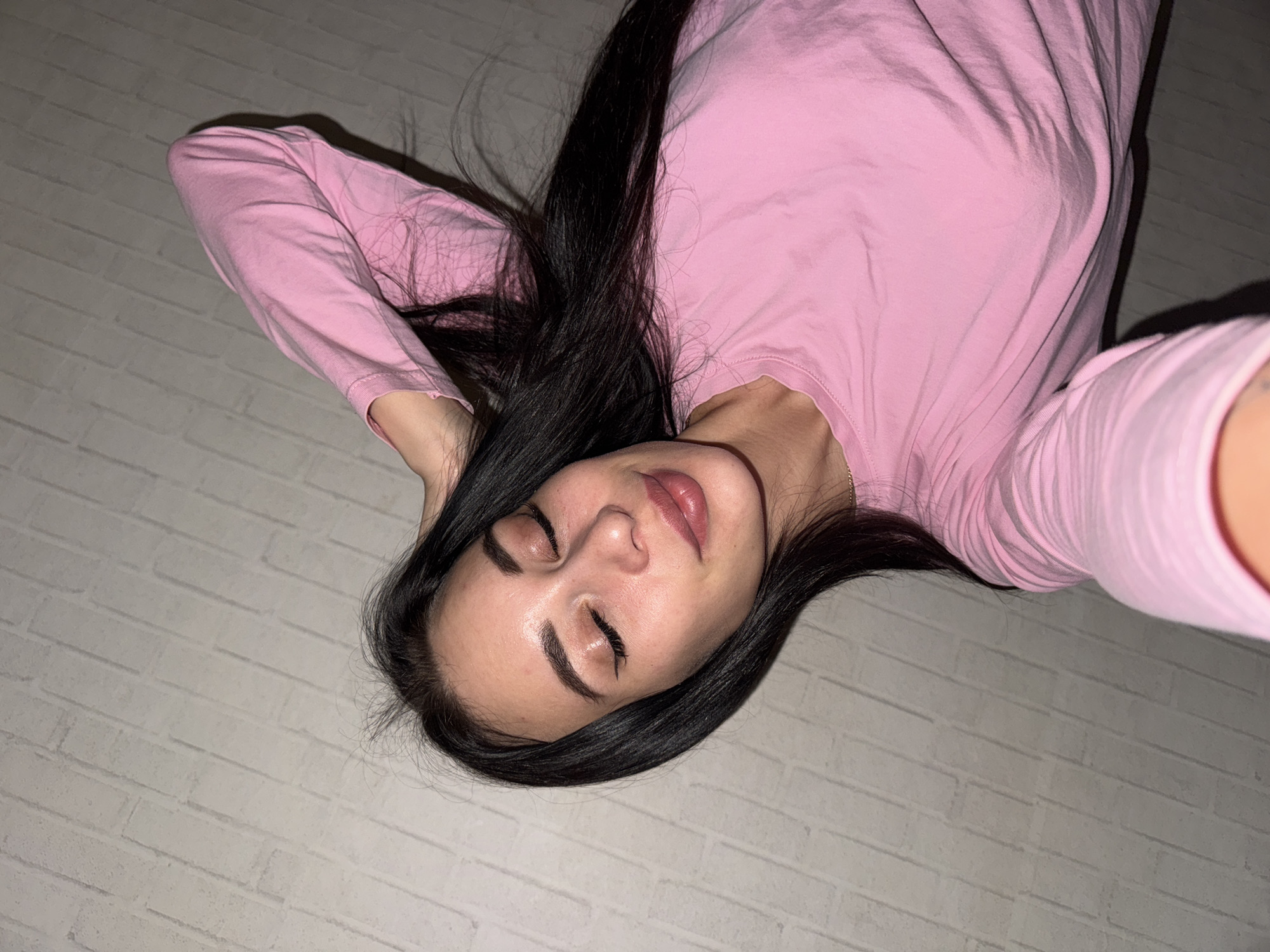 A woman with long, dark hair is lying down on a light-colored brick surface. She is wearing a pink long-sleeved shirt. Her eyes are closed. The camera is positioned above her at an angle. There is no evidence of the location being Chaikovsky, Russia.