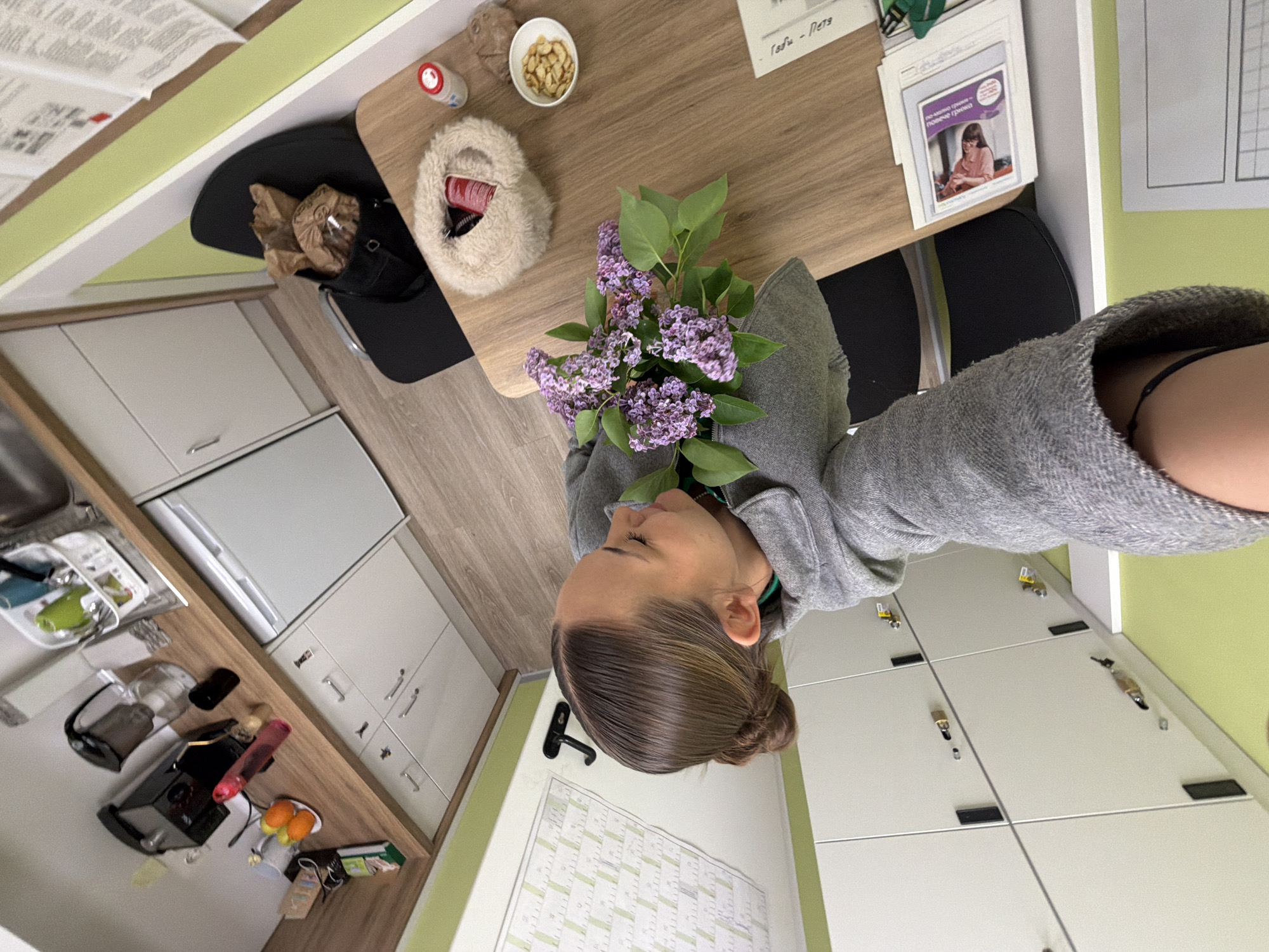 A woman in a gray hoodie is holding a bouquet of purple lilacs at a table. On the table, there is a small bowl of nuts, a small bottle, and a fluffy slipper. The background includes a kitchen area with white cabinets, a countertop with a coffee maker and fruit, a refrigerator, and a dining area with a table and chairs. Adjacent to the woman is a wall with a calendar and lockers. The specific location within Sofia, Bulgaria, is not ascertainable from this image.