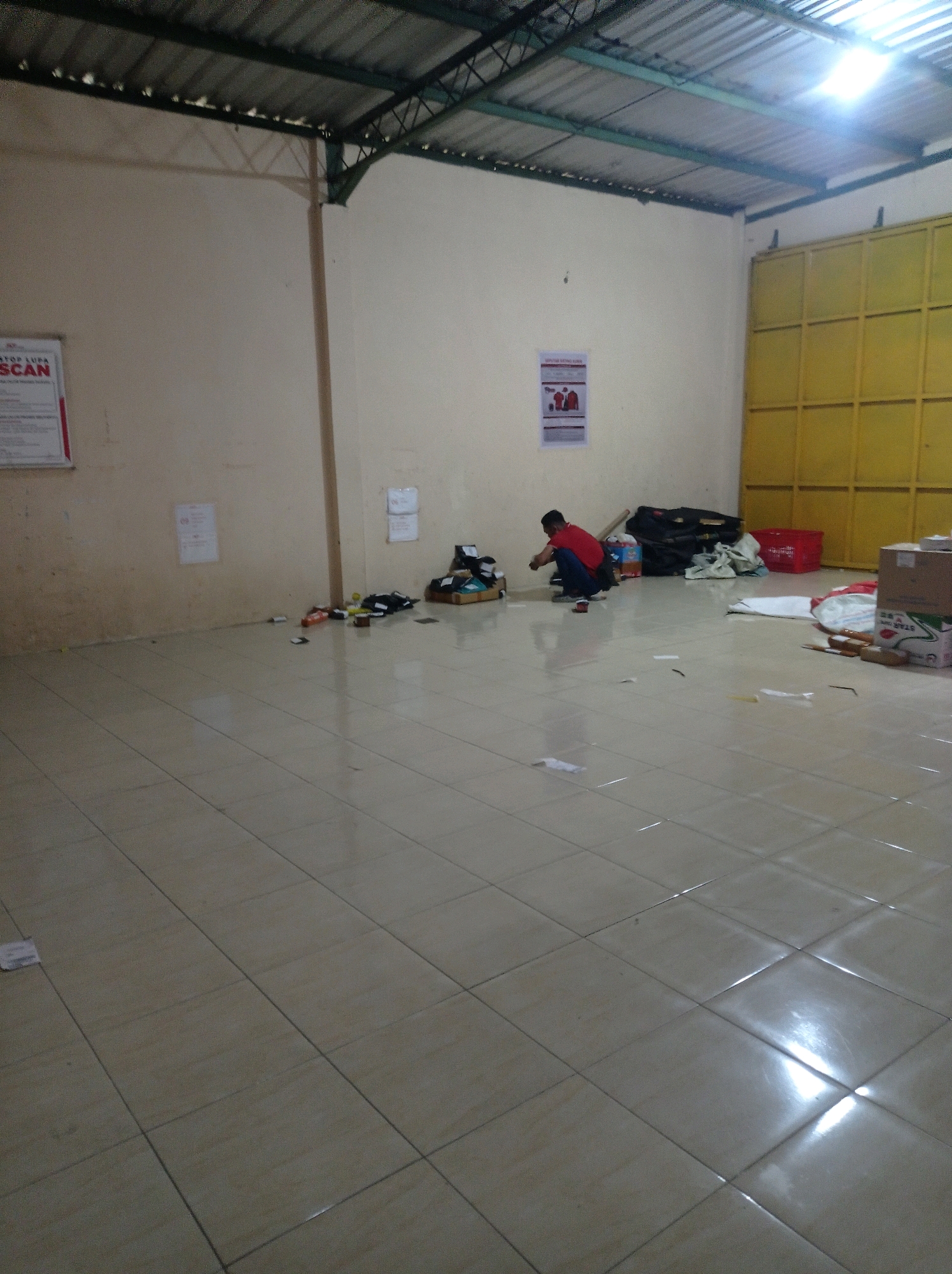 A man in a red shirt and dark pants is crouching on a tiled floor in what appears to be a warehouse or storage area in Jetis, Indonesia. The room has a corrugated metal roof supported by a metal frame and cream-colored walls. To the left, a poster is affixed to the wall. Further into the room, various items are scattered on the floor, including boxes, bags, and what appear to be some cleaning supplies. To the right, there is a large yellow gate or door. The floor is tiled and reflects the overhead lighting. The scene suggests a potentially untidy or in-progress work environment.