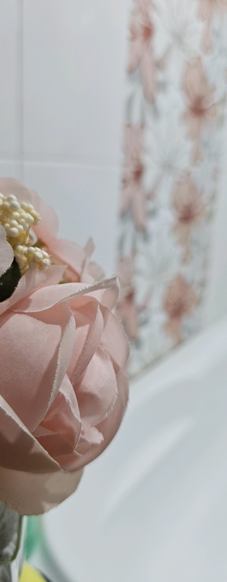 The image shows a close-up of a pale pink artificial rose with a cluster of small, light-colored buds. The rose is in focus in the foreground, with its delicate fabric petals visible. Behind the rose, out of focus, is a white surface with a subtle floral pattern in shades of pink and gray. To the right, a portion of a white, possibly ceramic or plastic, surface is visible, likely a sink or bathtub. The overall lighting is soft and diffused, suggesting an indoor environment. There are no people present, and no specific actions or interactions are depicted. The image's setting appears to be a bathroom, given the presence of a patterned shower curtain or wallpaper and a white fixture, which is consistent with a typical bathroom found in an apartment or house in Oskemen, Kazakhstan.