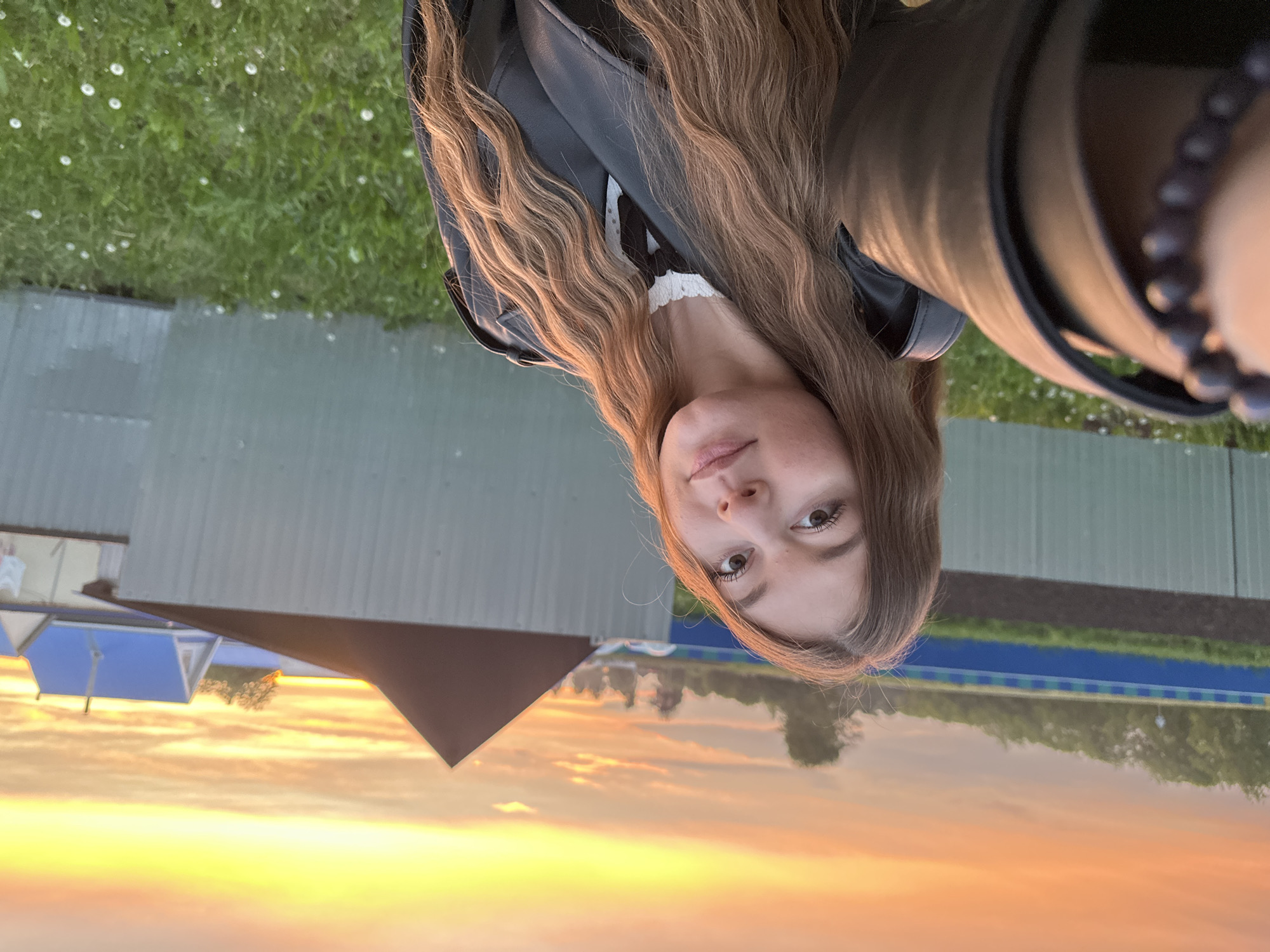 The image shows a selfie of a fair-skinned young woman with long, light brown hair wearing a black leather jacket. She has light eyebrows, brown eyes, and slightly parted lips. The background depicts an outdoor scene at sunset in Кузембетьево, Russia, featuring a fence, grass, and buildings. The sky is a gradient of orange, yellow, and pink hues. A reflective surface, likely water, creates a mirrored image of the sky and structures, adding to the surreal effect.